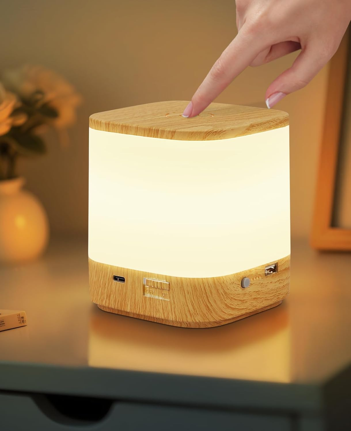 Una mano tocca una lampada di legno. La lampada emette una luce soffusa. È su un tavolo vicino a un vaso e una cornice.