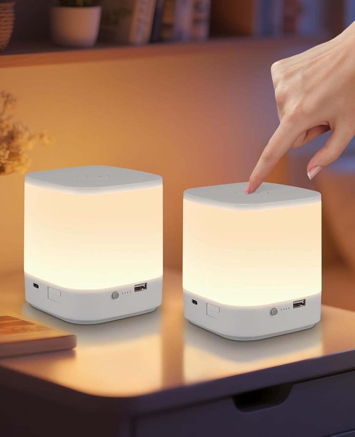 Due lampade a cubo bianche su un tavolo. Una mano tocca la parte superiore di una. Le lampade si illuminano delicatamente.