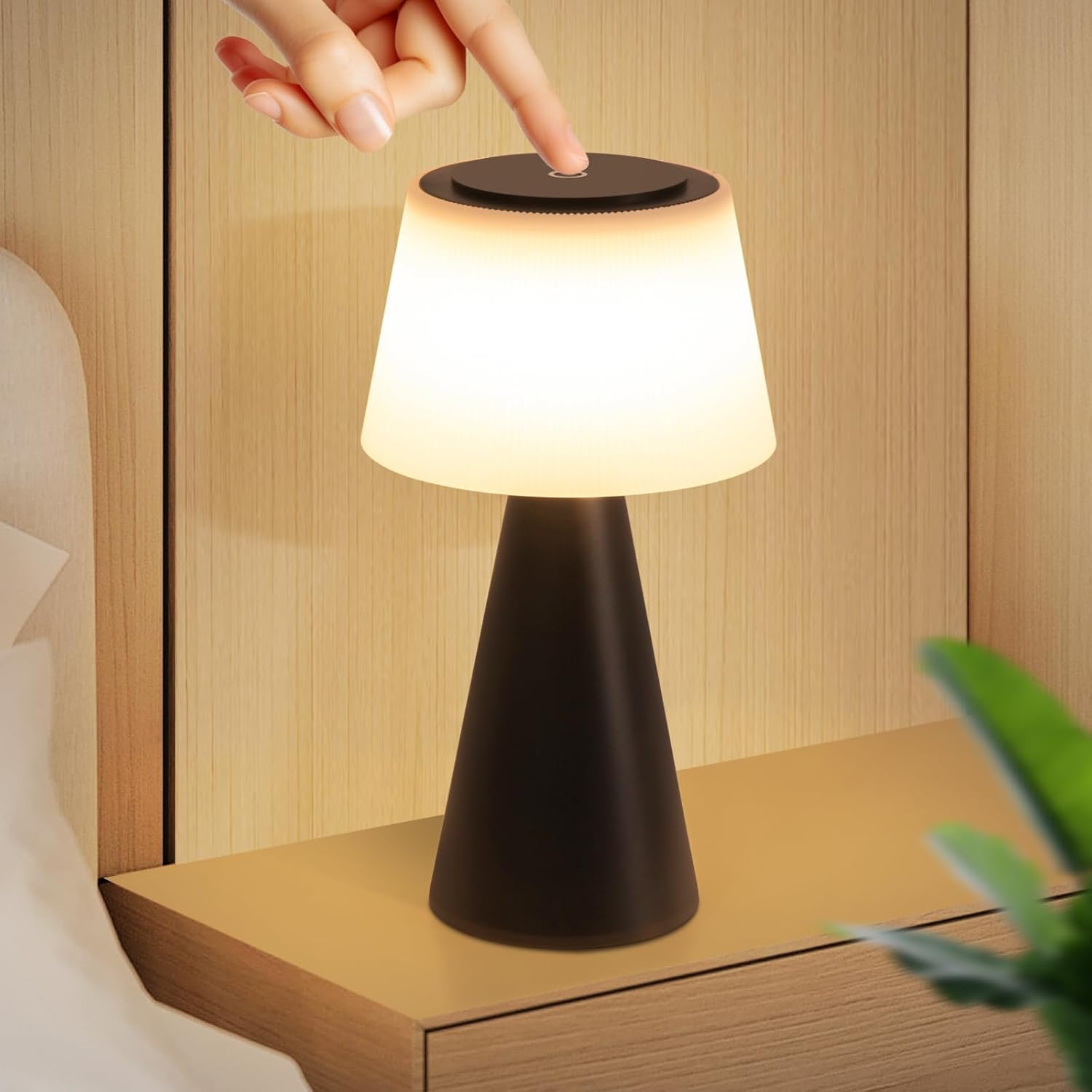 Una mano toca una lámpara negra sobre una mesita de noche. La lámpara está encendida, emitiendo un brillo cálido. Una planta al fondo.
