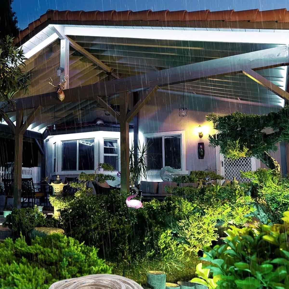 Eine regnerische Nachtszene einer beleuchteten Terrasse. Pflanzen und eine pinke Flamingo-Statue sind vorne. Ein Haus mit Lichtern ist hinten.