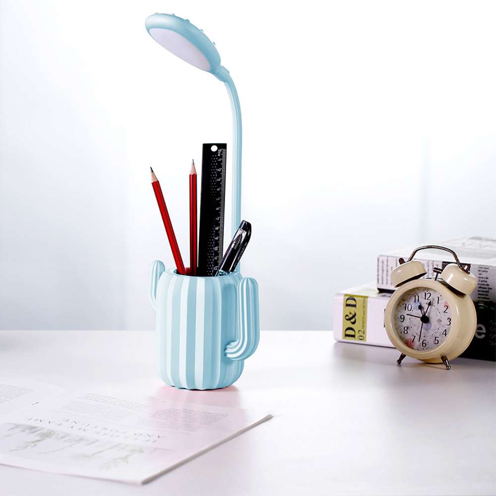 Eine Schreibtischszene mit einem kaktusförmigen Stifthalter, Bleistiften, einem Lineal, einer Lampe, einem Wecker und Büchern.