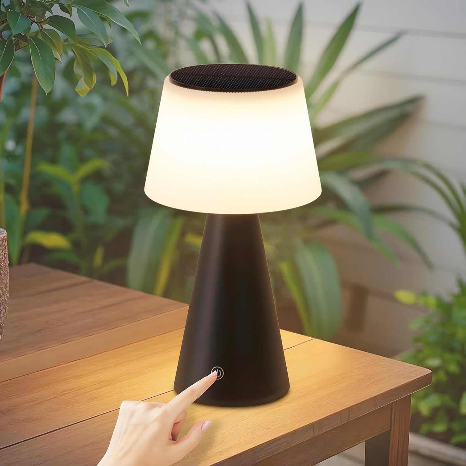 Una mano presiona el botón de encendido de una lámpara negra sobre una mesa de madera. La lámpara está encendida. Plantas verdes al fondo.