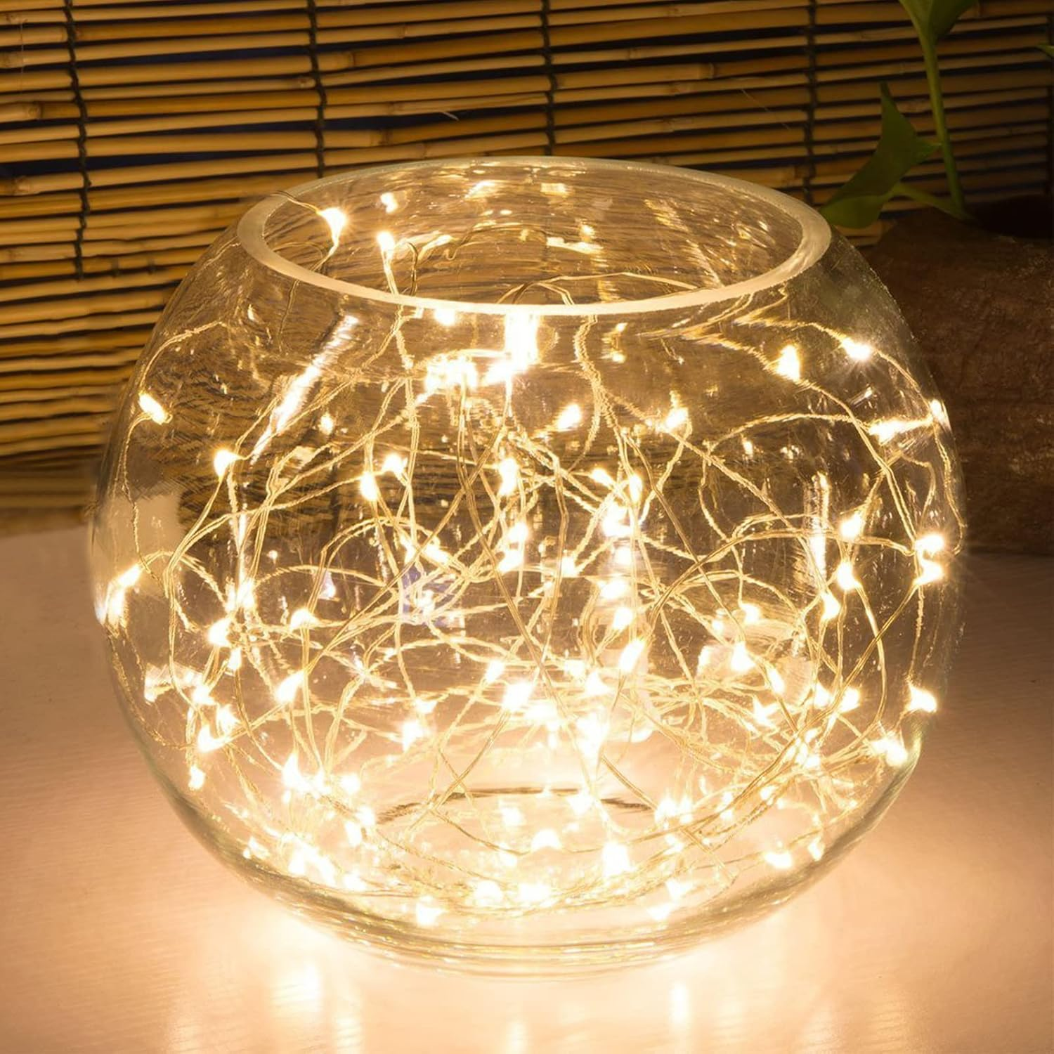 Eine klare Glasschale mit winzigen Lichterketten gefüllt. Im Hintergrund sind Bambusjalousien und Pflanzenblätter zu sehen.