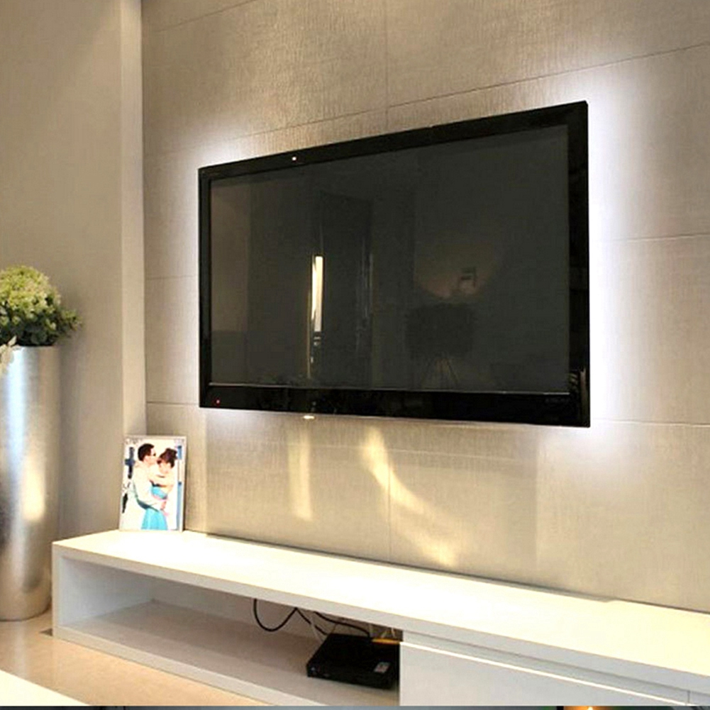 Ein großer schwarzer Fernseher hängt an einer hellgrauen Wand. Ein weißes Regal darunter beherbergt ein Foto und ein schwarzes Gerät.