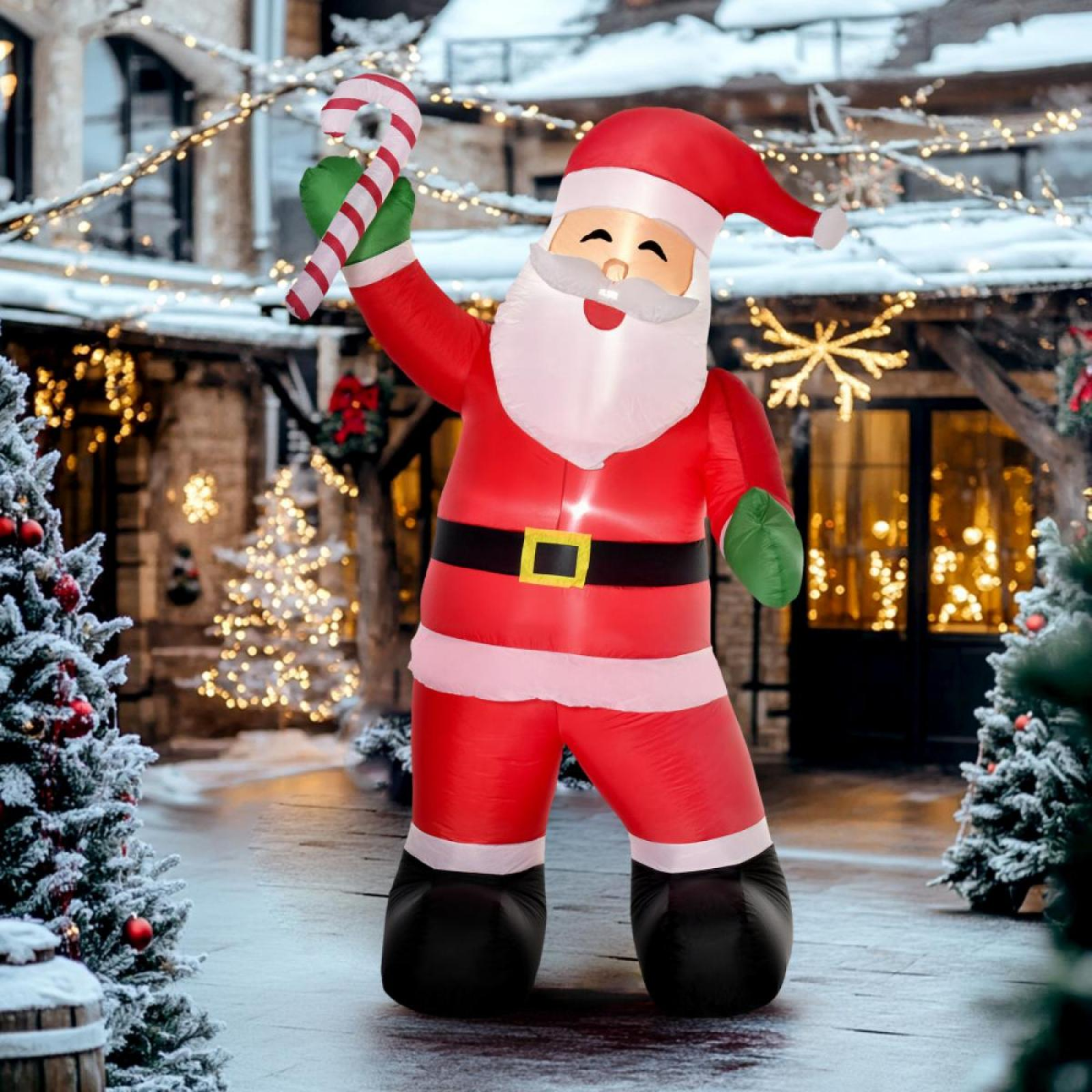 Aufblasbarer Weihnachtsmann mit Zuckerstange im Freien. Szene mit Weihnachtsbeleuchtung, Bäumen und verschneiter Umgebung.
