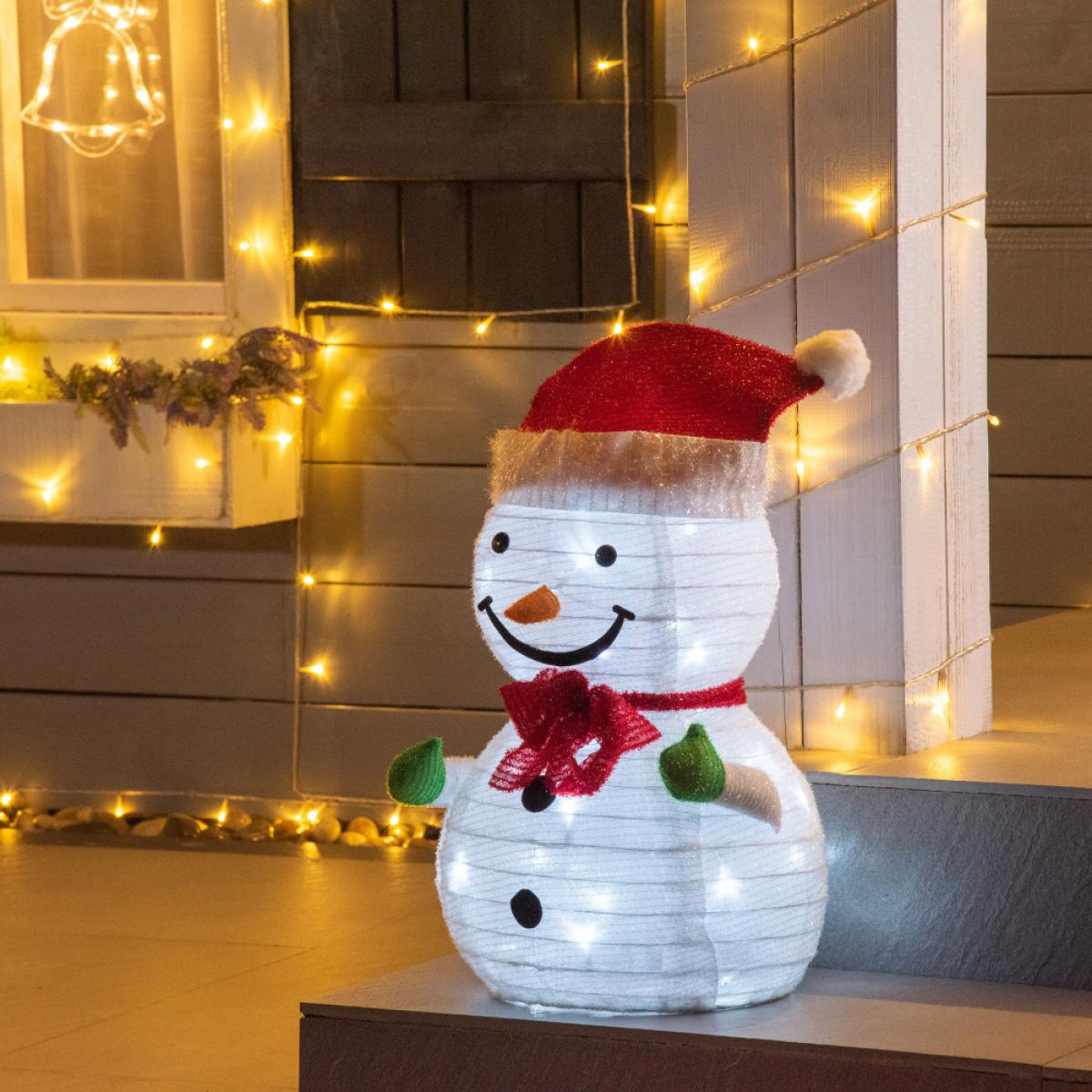 Eine Schneemann-Dekoration mit rotem Hut, Schal und grünen Fäustlingen. Er ist mit weißen Lichtern beleuchtet und steht auf einer Veranda mit Lichterketten.