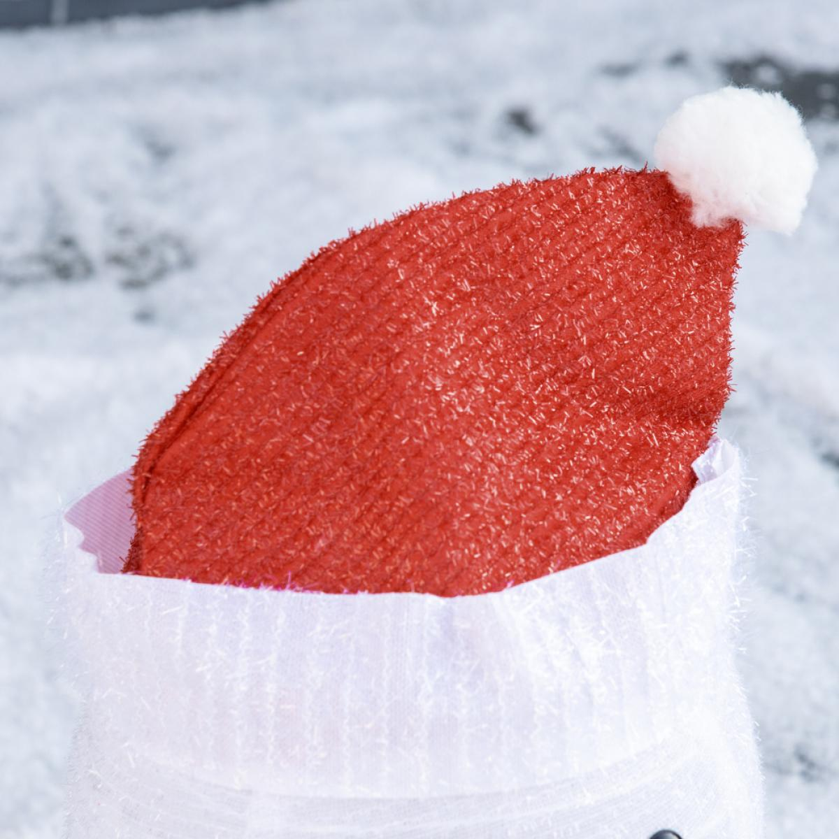 Ein roter Weihnachtsmannhut mit weißem Bommel liegt auf einem weißen Rand, vor verschwommenem, verschneitem Hintergrund.