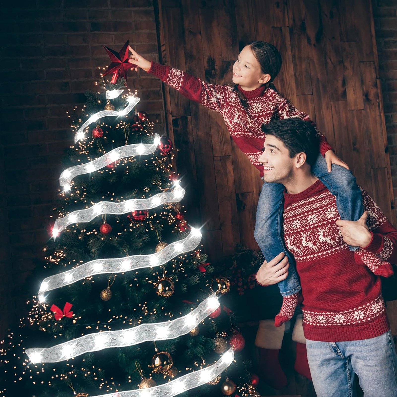Ein Vater trägt ein Kind auf seinen Schultern, um einen Stern auf eine Weihnachtstanne zu setzen. Beide tragen rote Pullover.