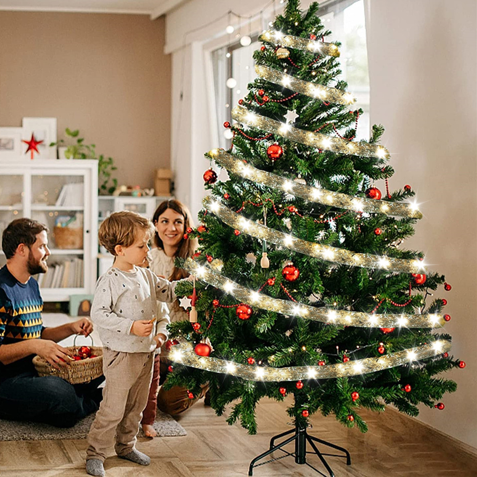 Eine Familie schmückt einen Weihnachtsbaum mit roten Ornamenten und Lichtern. Ein Kind und Eltern sind anwesend.