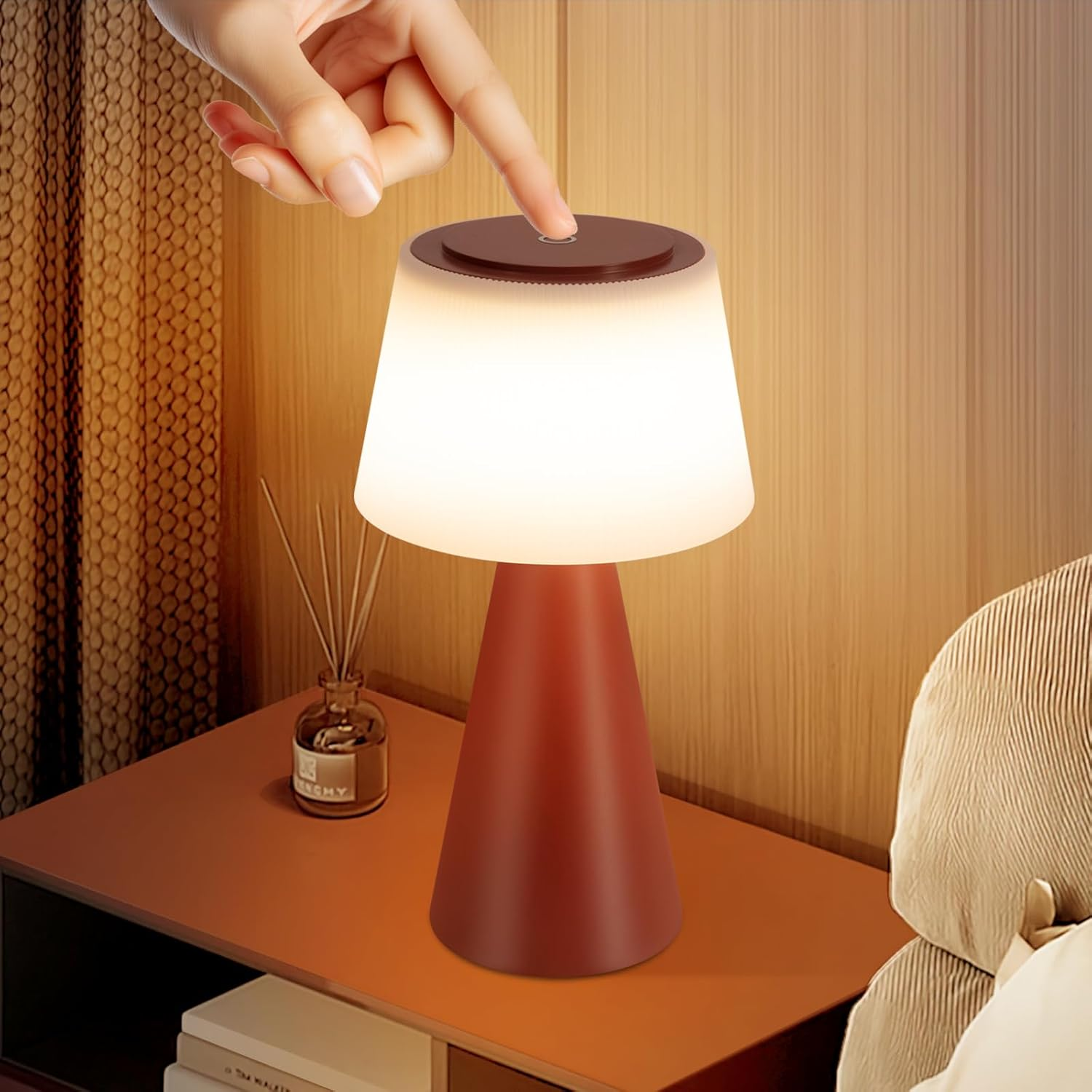 Una mano tocca una lampada. La lampada è su un tavolo con un diffusore e libri. Lo sfondo include un muro e un letto.