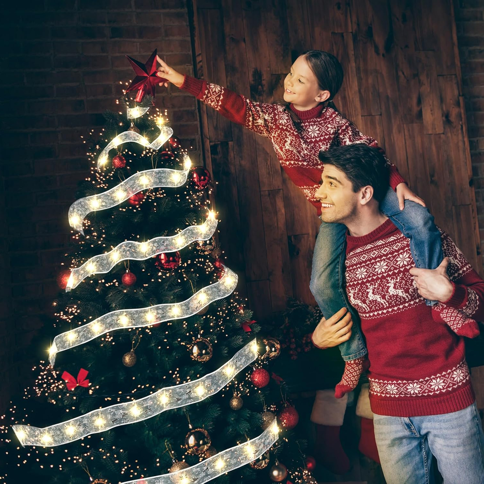 Vater trägt Tochter auf Schultern, um einen Stern auf den Weihnachtsbaum zu setzen. Beide tragen rote Pullover.
