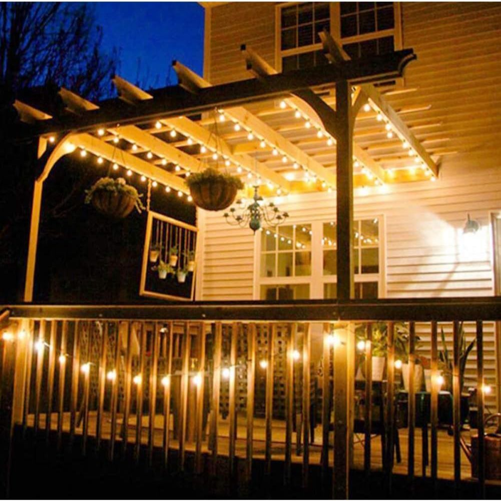 Eine Holzterrasse mit Lichterketten, Hängepflanzen und einem Kronleuchter. Vorne ein Holzzaun, im Hintergrund ein Haus.