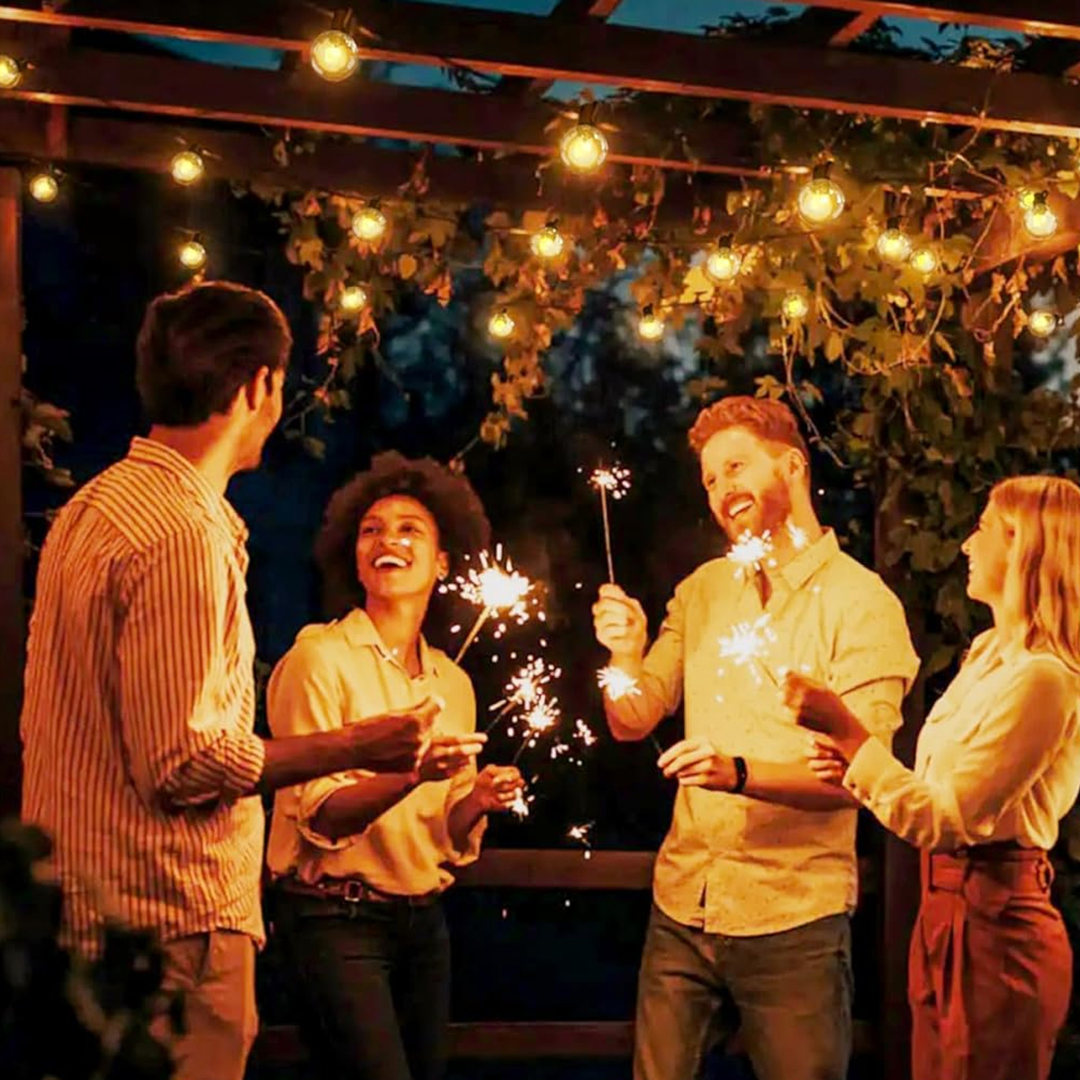 Vier Personen feiern nachts mit Wunderkerzen unter Lichterketten. Sie lächeln und lachen.