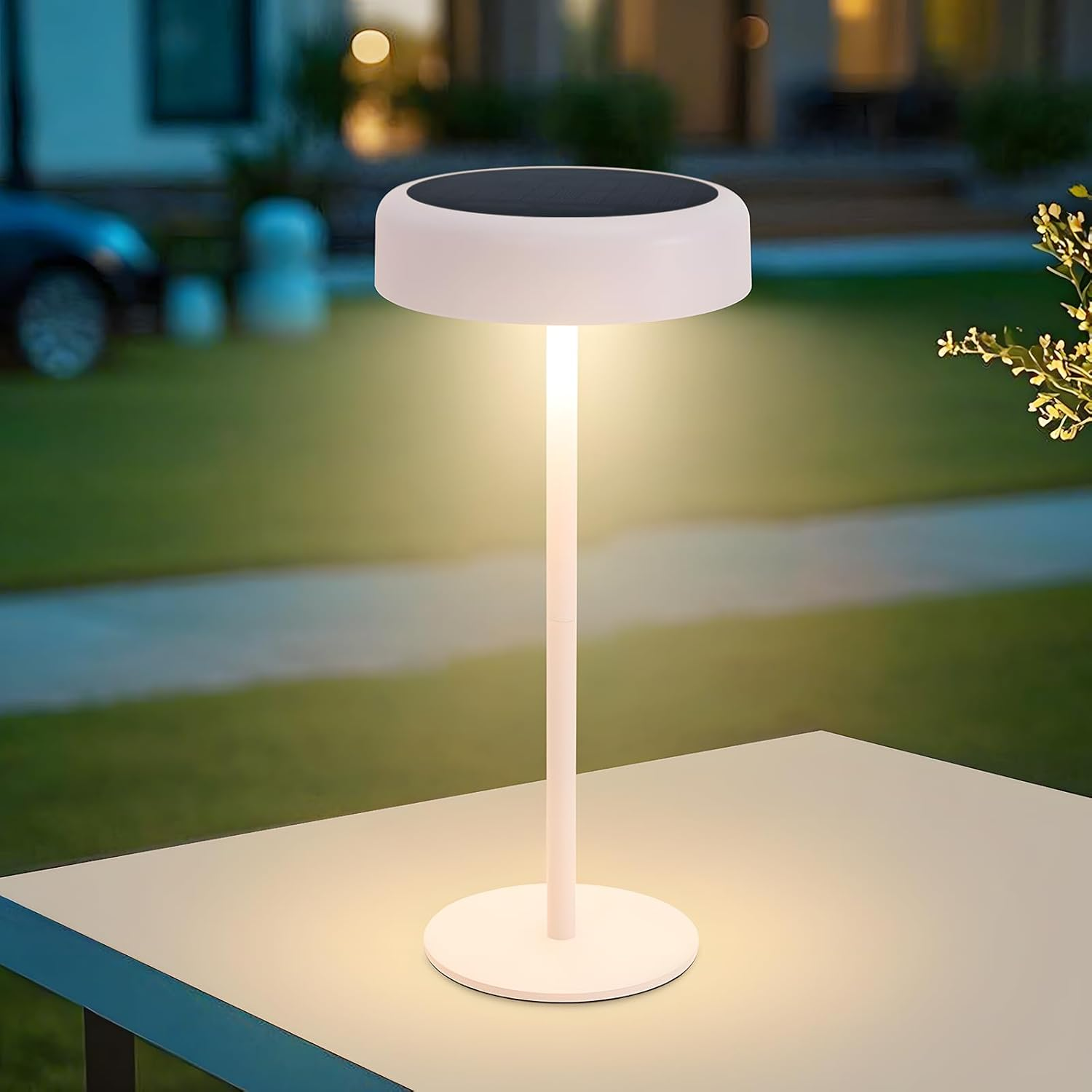 Una lampada bianca a energia solare è su un tavolo. Ha una base rotonda, uno stelo e una parte superiore con pannello solare. Lo sfondo è un giardino.