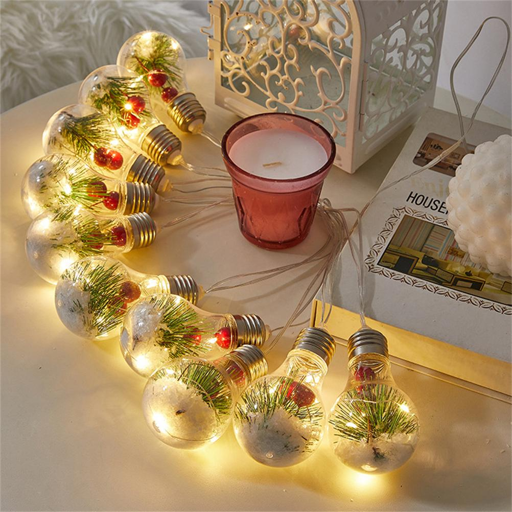 Eine Lichterkette mit glühbirnenförmigen Lichtern, jede mit kleinen Pflanzen und roten Beeren. Eine Kerze und ein Buch sind in der Nähe.