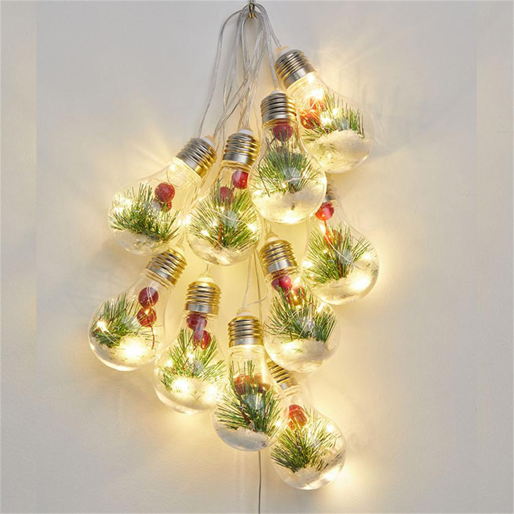 Lichterkette mit dekorativen Glühbirnen. Jede Birne enthält Grün, Beeren und kleine Lichter. Sie hängen an einer weißen Wand.
