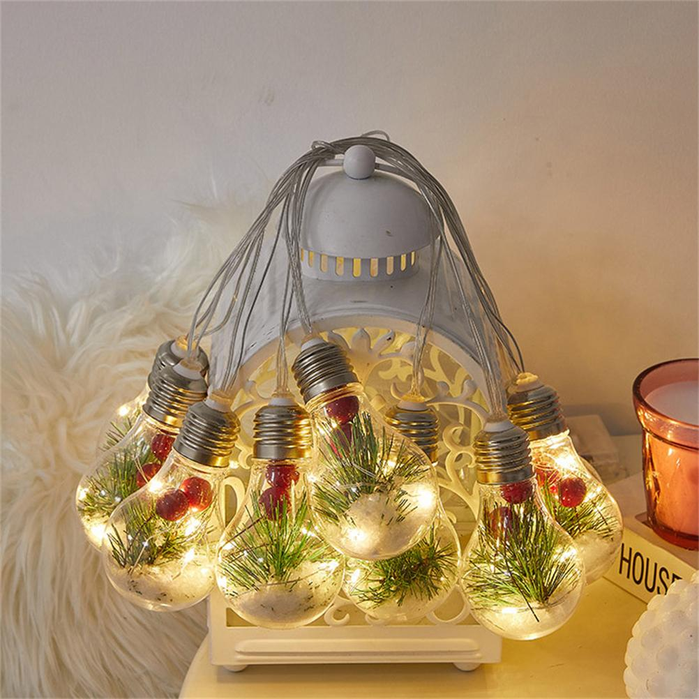 Lichterketten mit Glühbirnen, gefüllt mit grünen Zweigen und roten Beeren. Sie hängen vor einer weißen dekorativen Laterne.