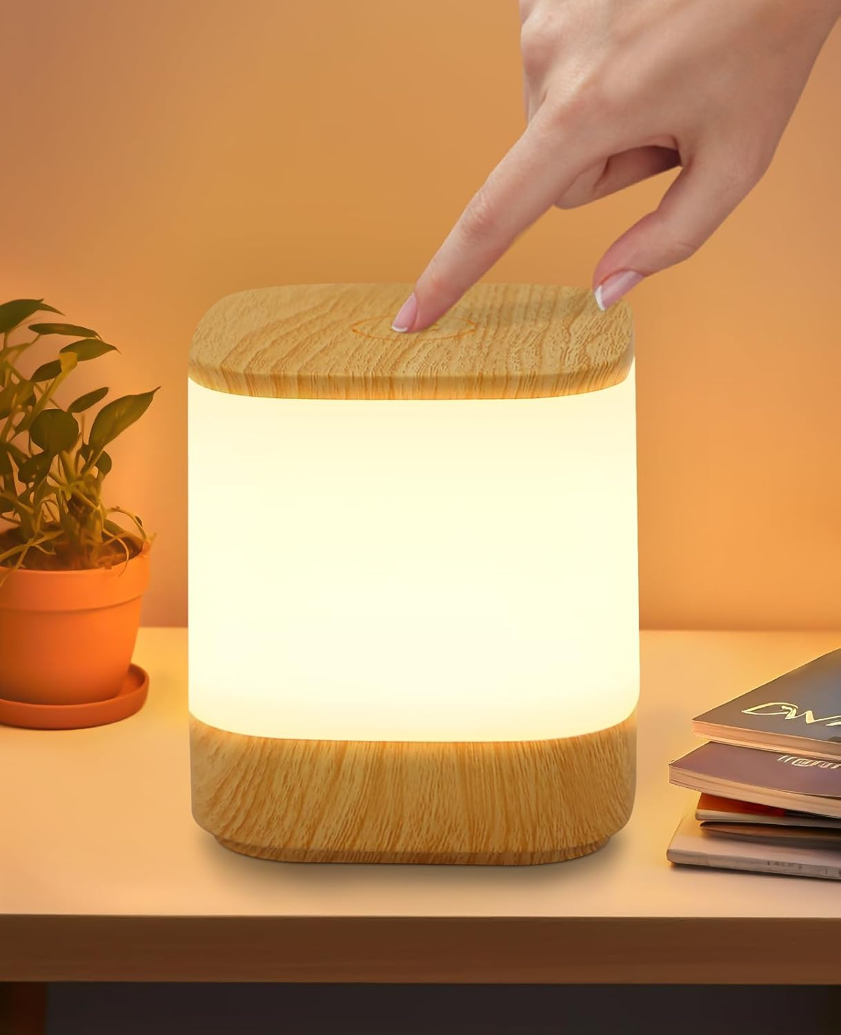 Una mano tocca una lampada con la parte superiore in legno che emette una luce soffusa. Una pianta in vaso e libri sono sul tavolo. Lo sfondo è sfumato.