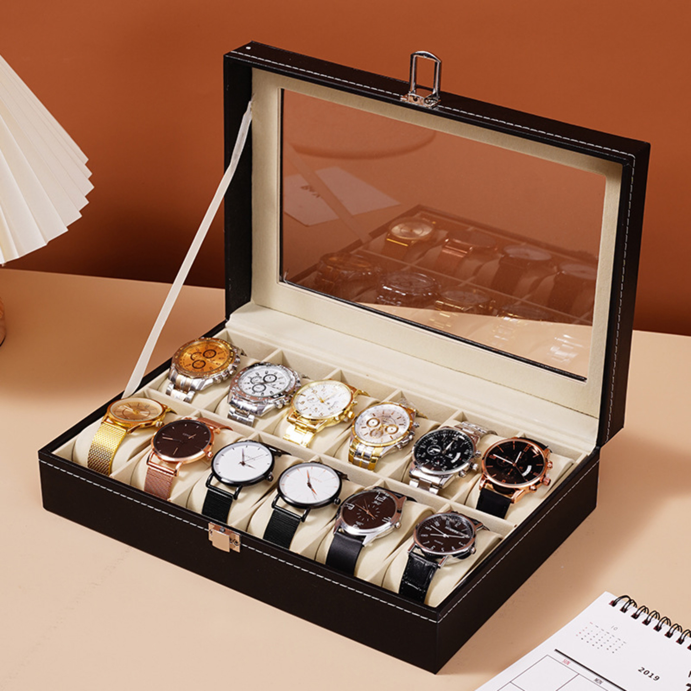 Eine schwarze Uhrenbox mit 12 Uhren. Links ist eine Lampe. Rechts ist ein Kalender.