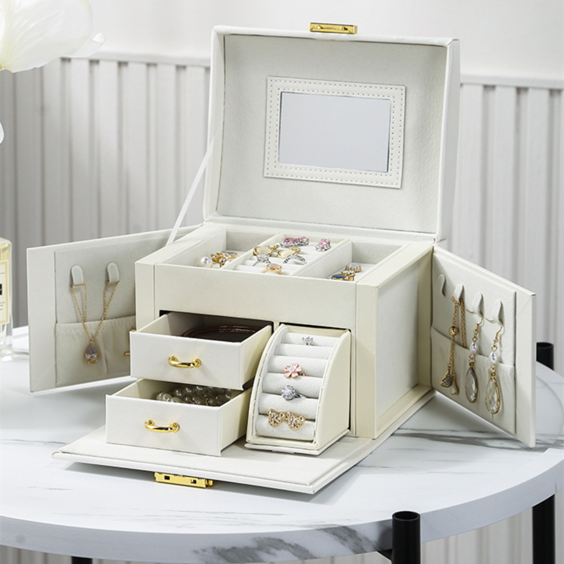 Geöffnete cremefarbene Schmuckschatulle mit Spiegel, Schubladen und Schmuck. Goldene Beschläge. Ringe, Halsketten und Ohrringe sind sichtbar.