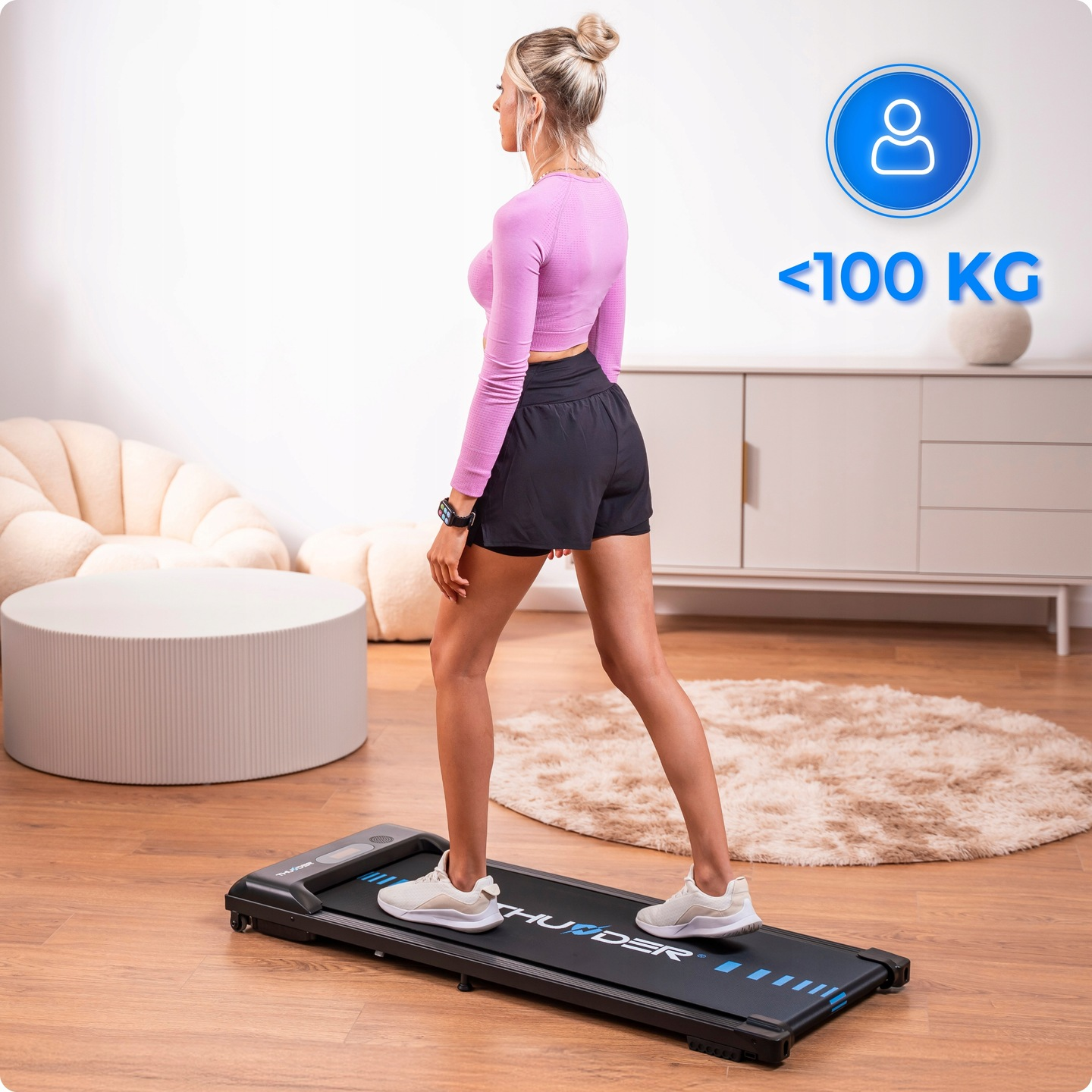 Kobieta ćwiczy na bieżni. Ma na sobie szorty i różowy top. Znak wskazuje, że limit wagi wynosi mniej niż 100 kg.