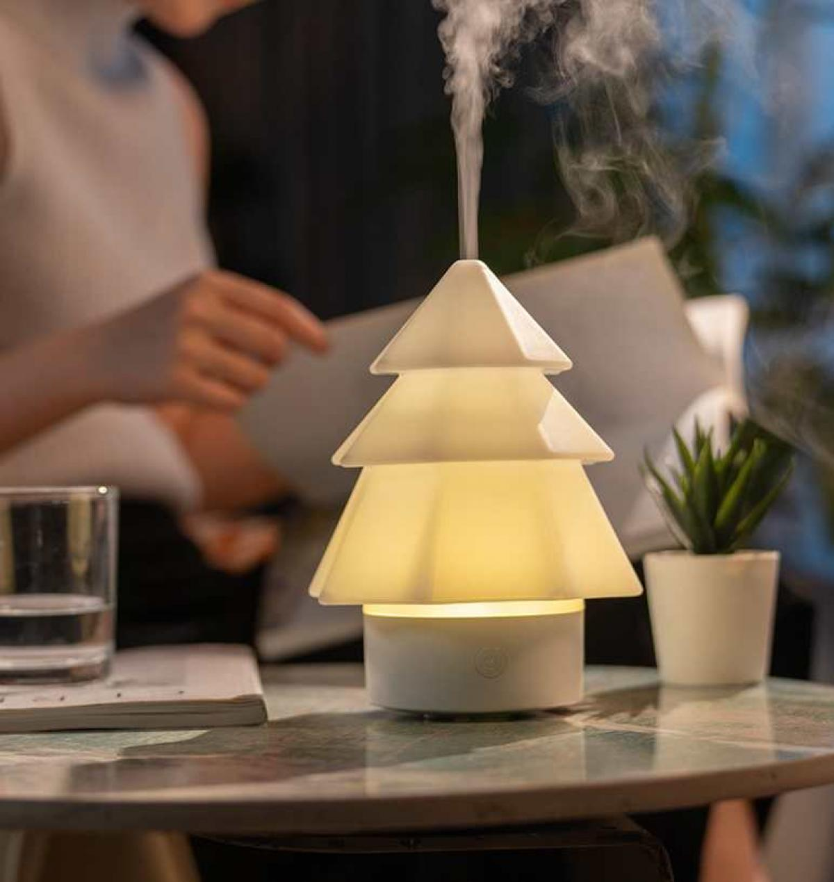 Ein weißer, weihnachtsbaumförmiger Luftbefeuchter gibt Dampf ab. Im Hintergrund liest eine Person. Ein Glas, eine Pflanze und ein Notizbuch stehen auf dem Tisch.