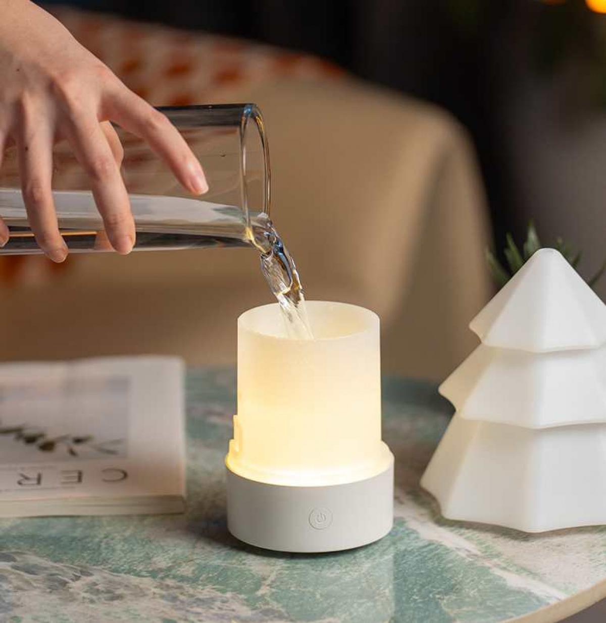 Eine Hand gießt Wasser aus einem Glas in einen leuchtenden weißen Zylinder. Rechts befindet sich eine weiße Baumdekoration. Links liegt ein Buch.