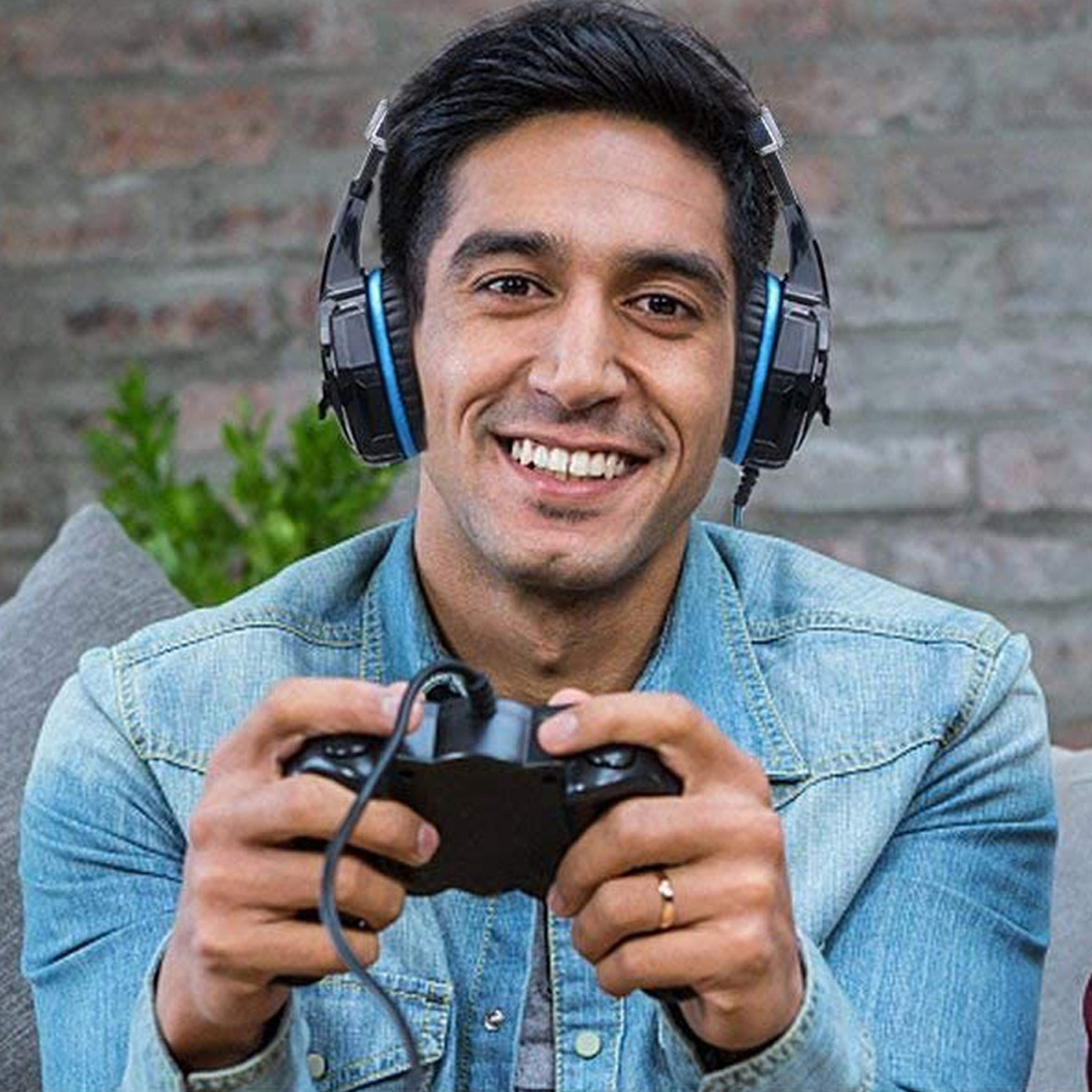Ein Mann lächelt, trägt Kopfhörer und hält einen Gamecontroller. Er trägt eine Jeansjacke, Ring am Finger. Hintergrund: Backsteinmauer.