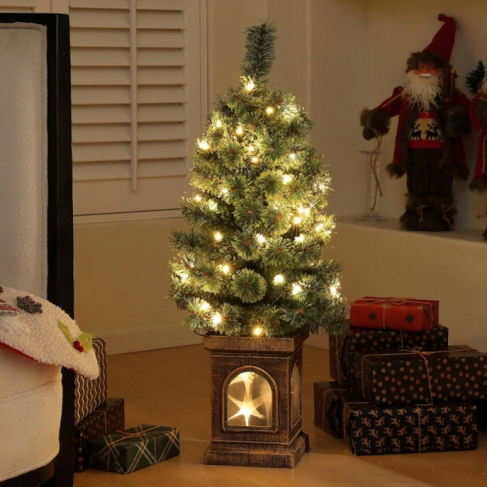 Ein kleiner Weihnachtsbaum mit Lichtern. Geschenke sind daneben gestapelt. Eine Santa-Figur steht im Hintergrund.