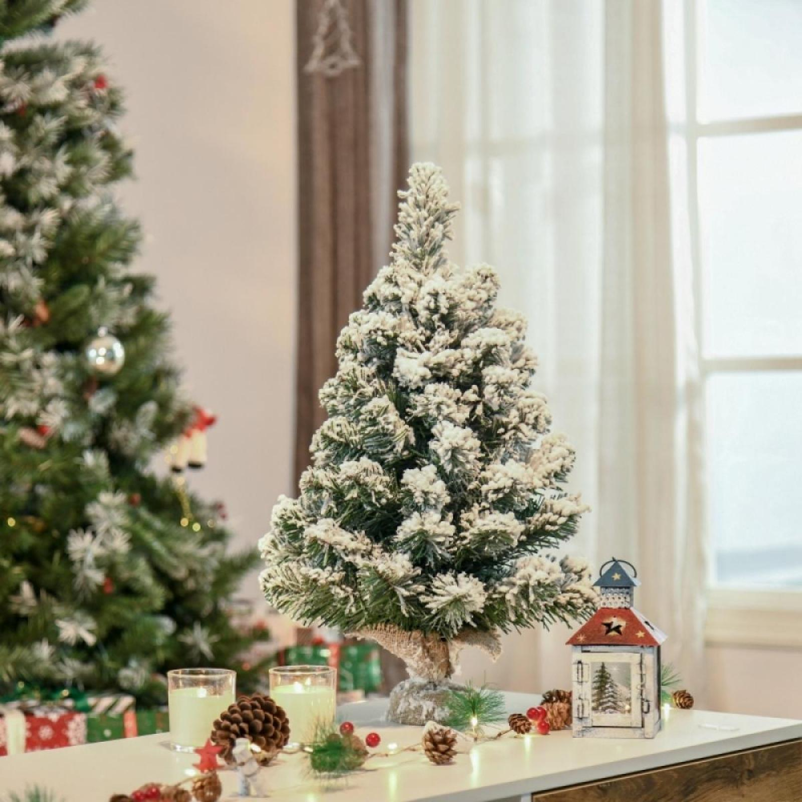 Ein kleiner Weihnachtsbaum mit Schnee, eine Laterne, Kerzen und Tannenzapfen auf einem Tisch. Ein anderer Baum im Hintergrund.