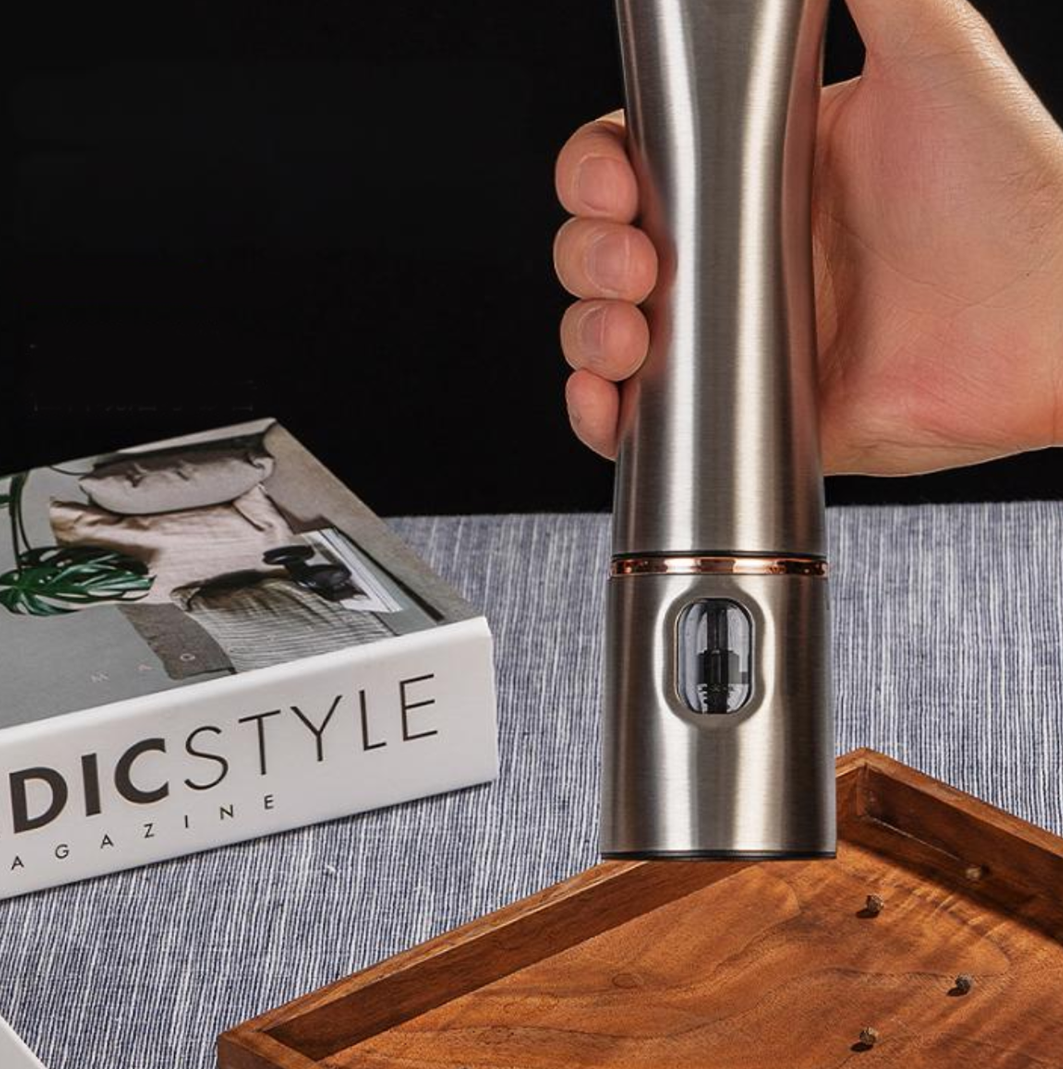 Eine Hand hält eine silberne elektrische Pfeffermühle. Ein Magazin und ein Holztablett sind ebenfalls zu sehen.