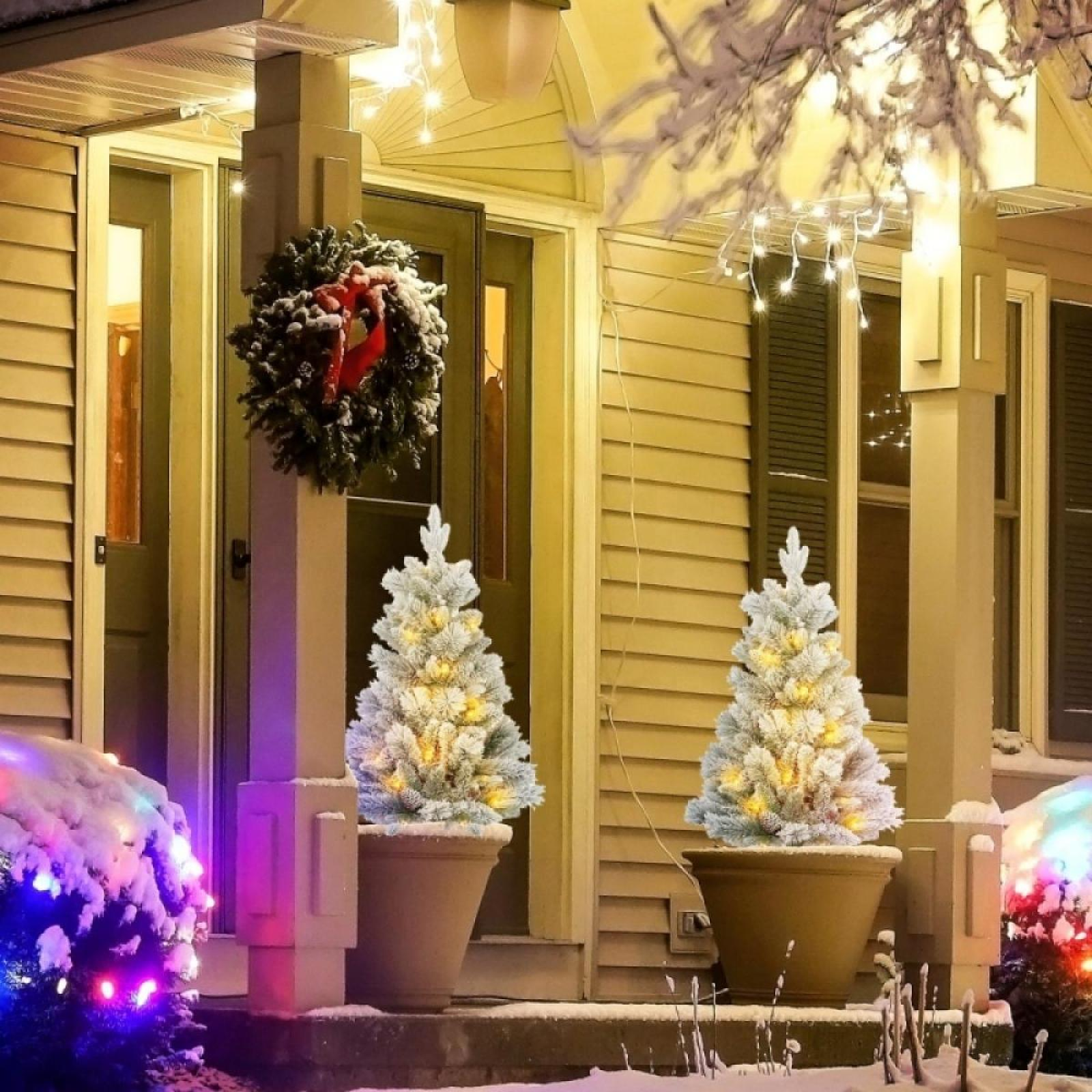 Eine verschneite Veranda mit zwei beleuchteten Weihnachtsbäumen in Töpfen. Ein Kranz hängt an der Tür. Lichterketten und Schnee auf den Büschen.