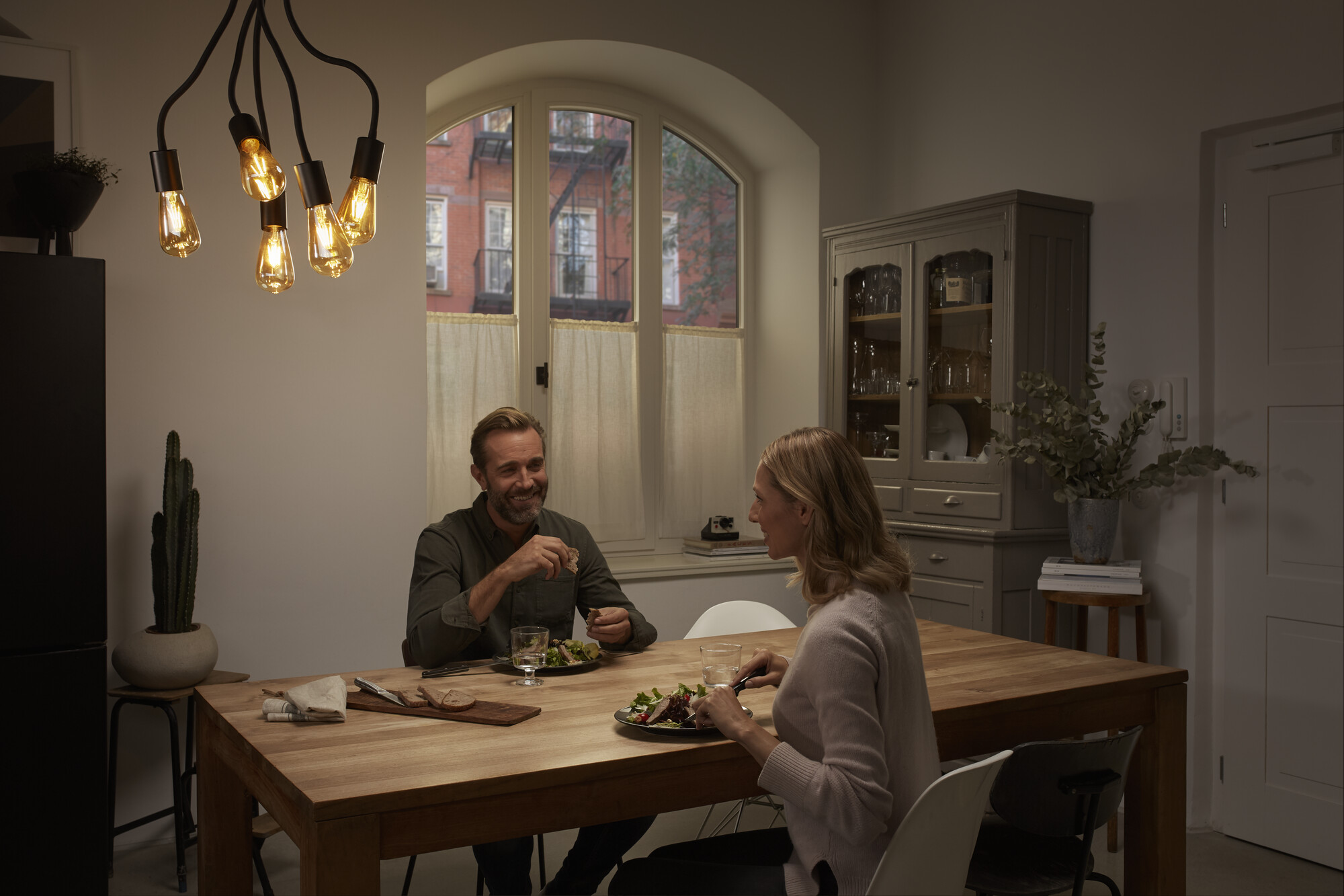 Para je obiad przy drewnianym stole. Żyrandol wisi nad nimi. Tło przedstawia okno z widokiem na miasto.