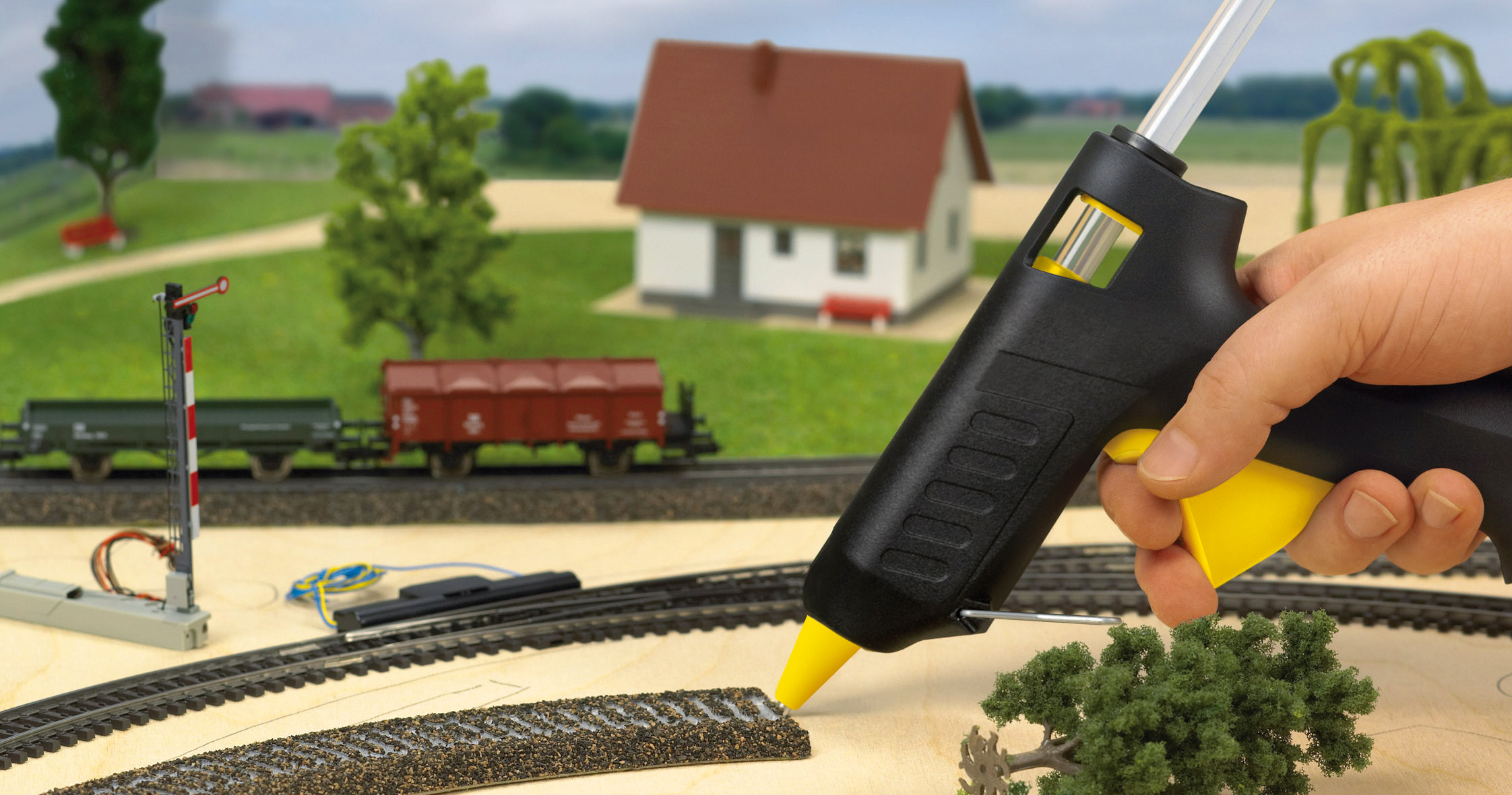 Eine Hand hält eine Heißklebepistole und klebt eine Miniatur-Eisenbahnstrecke. Ein Haus, Züge und Bäume sind im Hintergrund.