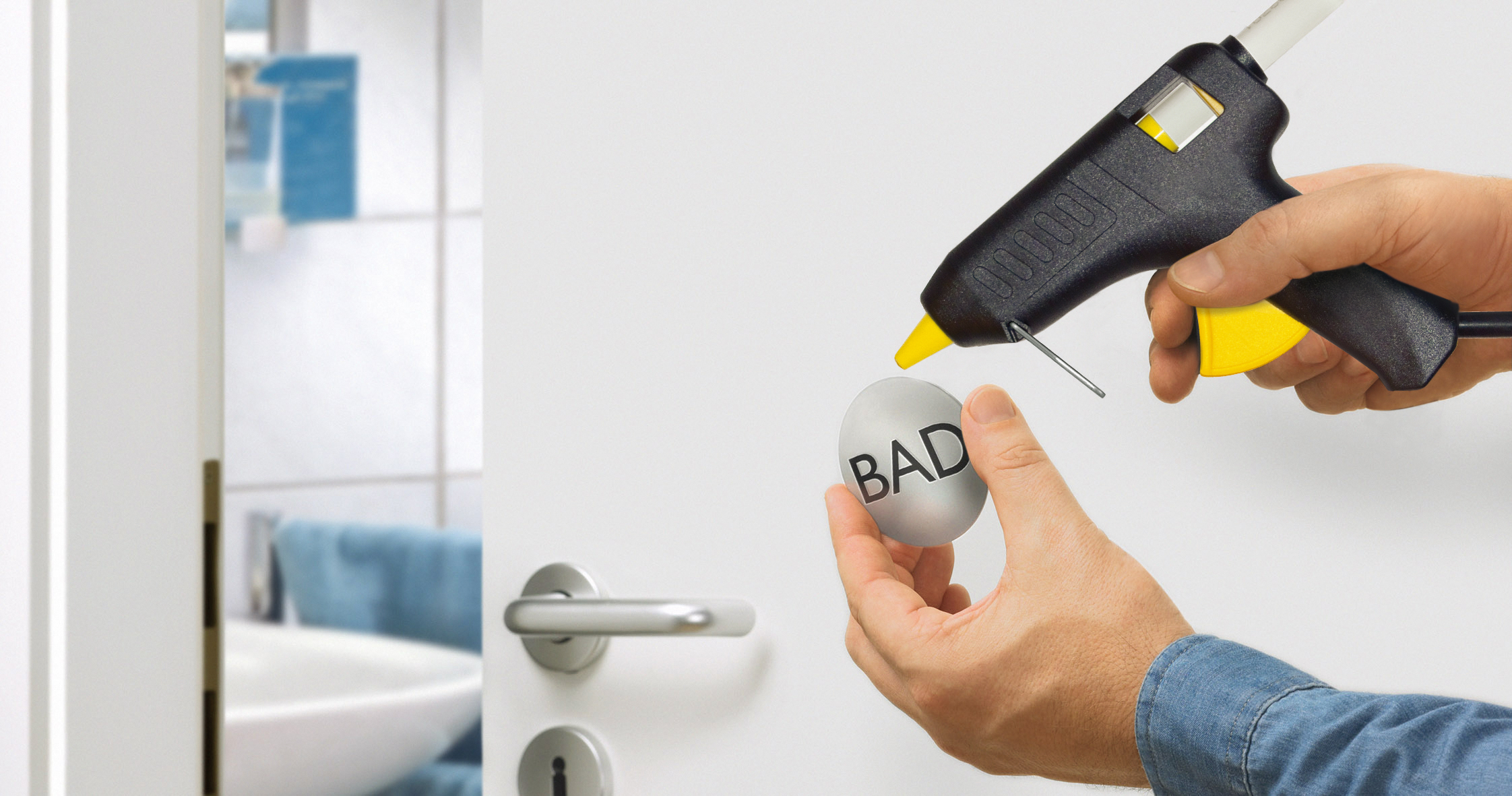 Eine Hand hält eine Heißklebepistole über einer silbernen Kugel mit der Aufschrift 'BAD' in der Nähe einer weißen Tür.