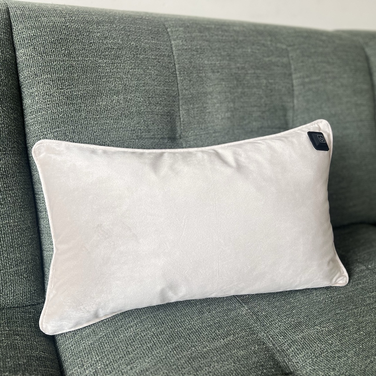 Ein weißes rechteckiges Kissen mit einem Power-Knopf liegt auf einem grünen Sofa.