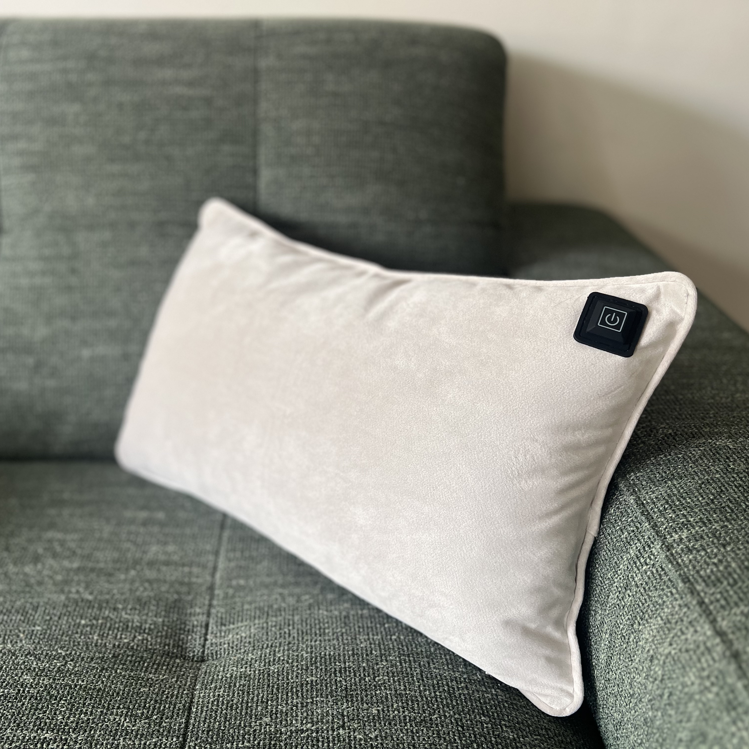 Ein hellgraues Kissen mit schwarzem Power-Knopf auf einer grün strukturierten Couch. Hintergrund ist eine Wand.