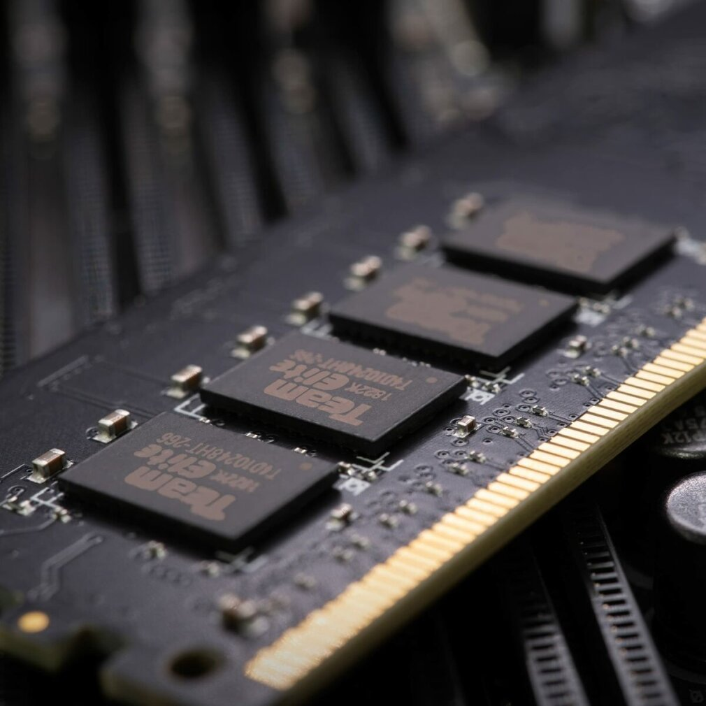 Primo piano di un modulo RAM nero con contatti dorati e quattro chip quadrati con testo. Lo sfondo è sfocato.