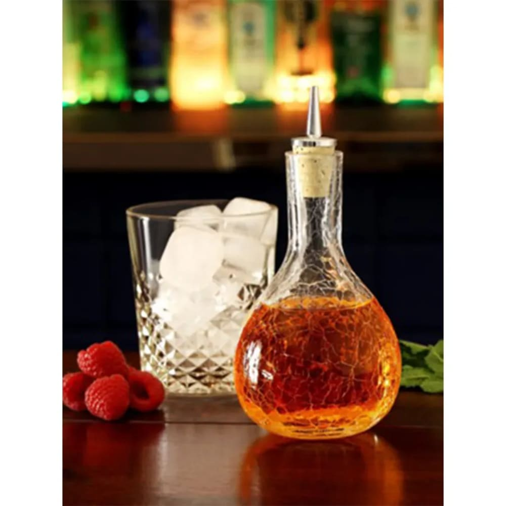 Una caraffa di vetro con liquido ambrato, un bicchiere di cubetti di ghiaccio e lamponi su una superficie di legno, con sfondo bar.