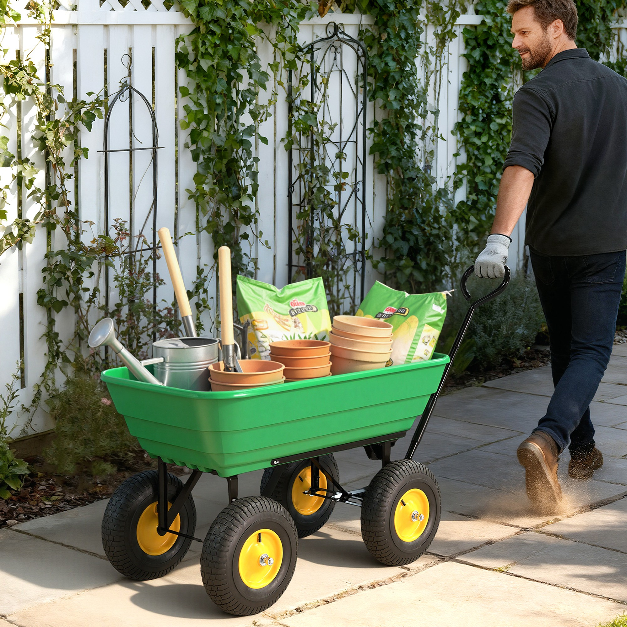 Un uomo tira un carrello verde con attrezzi da giardinaggio, terra e vasi. Edera e staccionata bianca sullo sfondo.