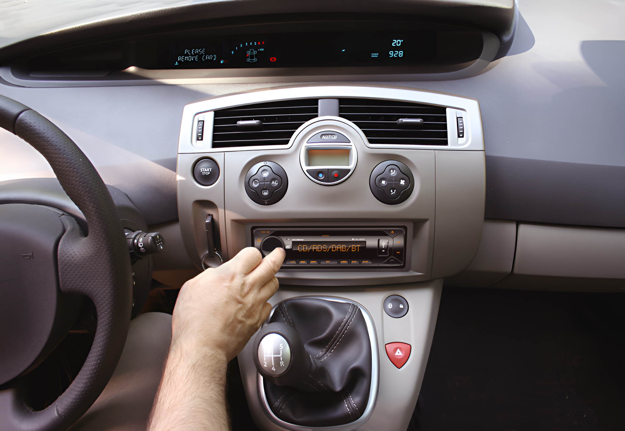 Car interior. Hand operating the radio. Gear stick, steering wheel, and dashboard are visible. Temperature display shows 20 degrees.