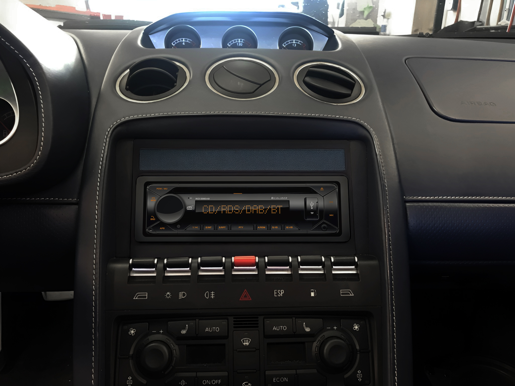 Car dashboard showing a radio, air vents, and gauges. Black and blue interior with white stitching. Red hazard light button is visible.