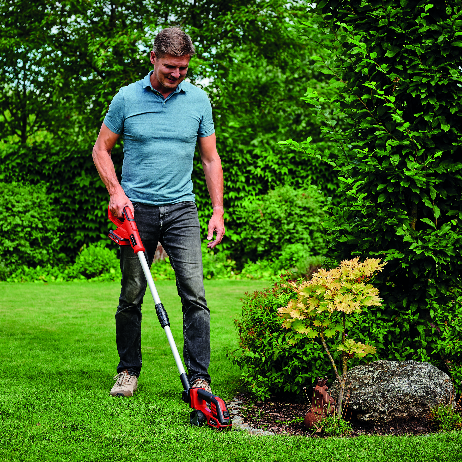 Ein Mann benutzt einen roten und silbernen Gartenschneider in einem grünen Garten, in der Nähe von Büschen und Bäumen.