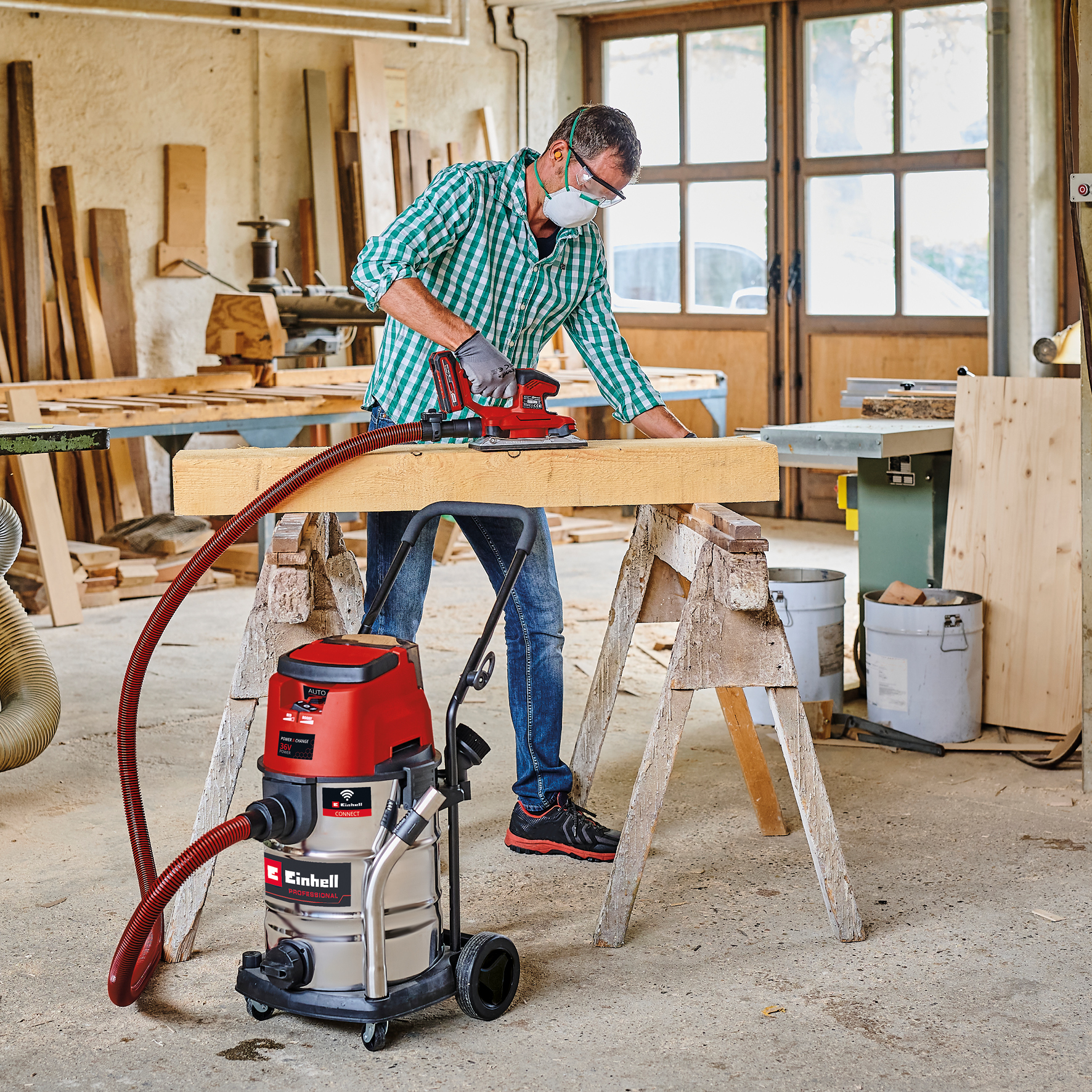 Ein Mann schleift Holz mit einem roten Schleifgerät. Ein Staubsauger ist angeschlossen. Werkstatteinstellung mit Werkzeugen und Holz.