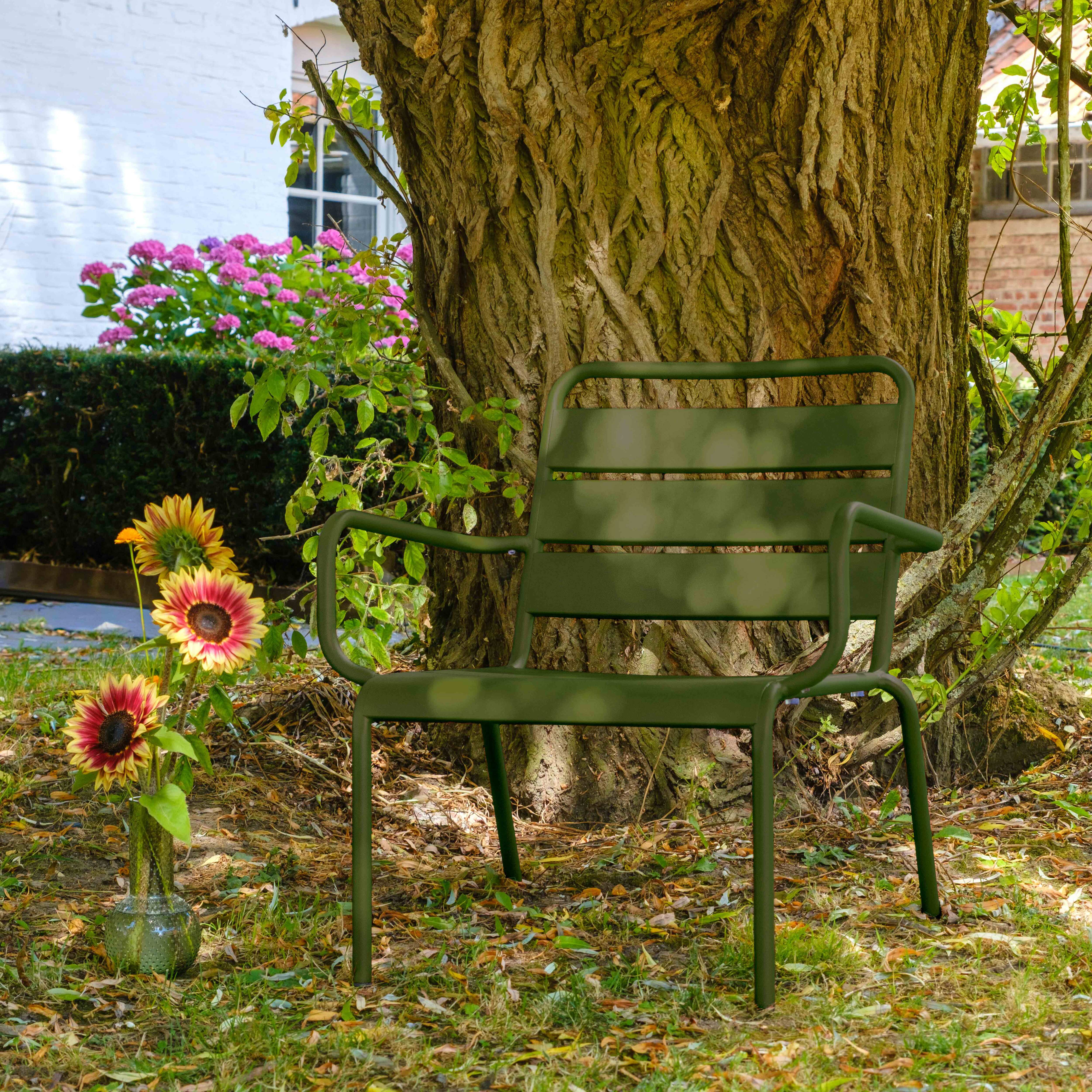 Grüner Stuhl neben einem Baumstamm. Sonnenblumen in einer Vase links. Rosa Blumen und ein weißes Gebäude im Hintergrund.