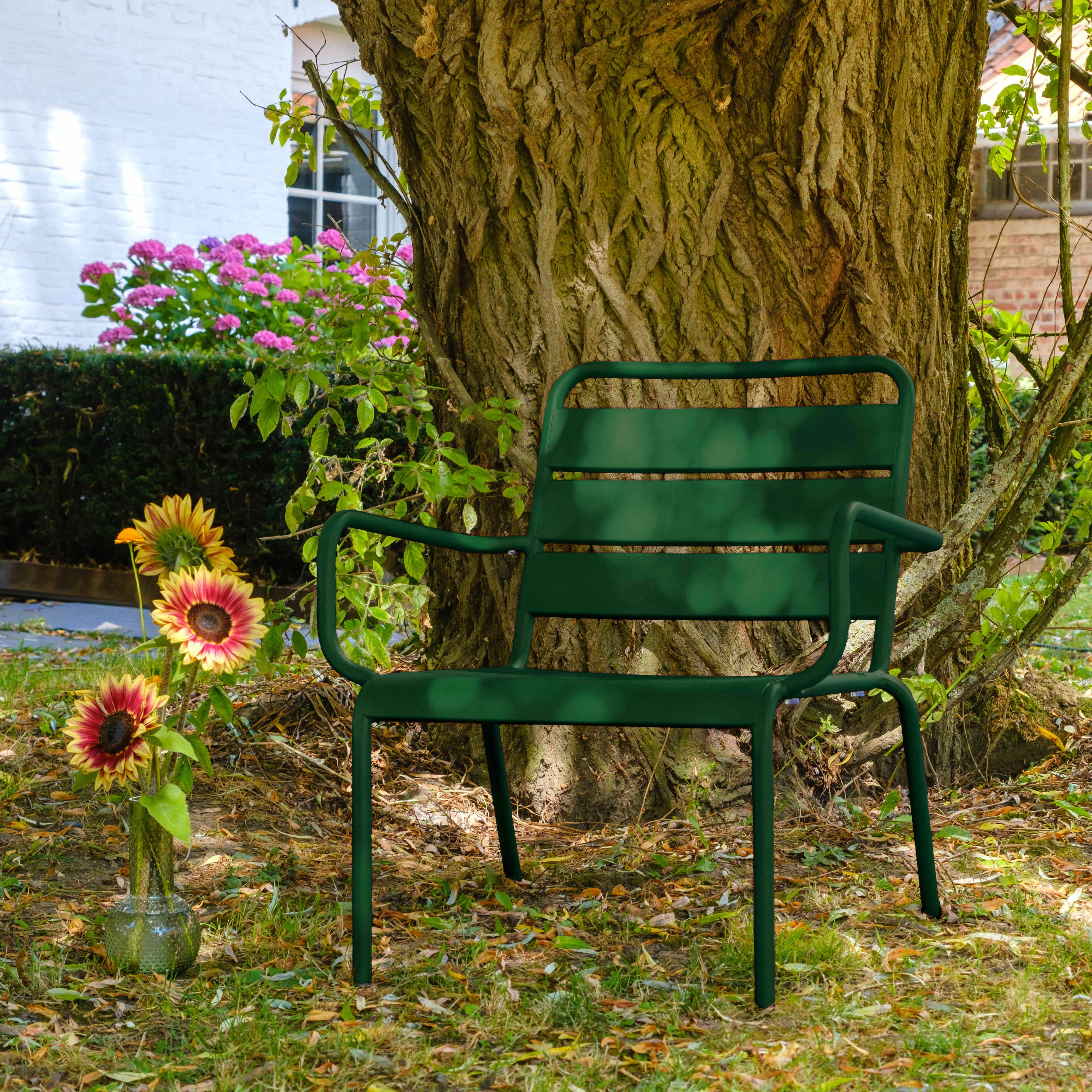 Ein grüner Stuhl steht unter einem großen Baum. Sonnenblumen stehen in einer Vase. Im Hintergrund ist ein weißes Gebäude.