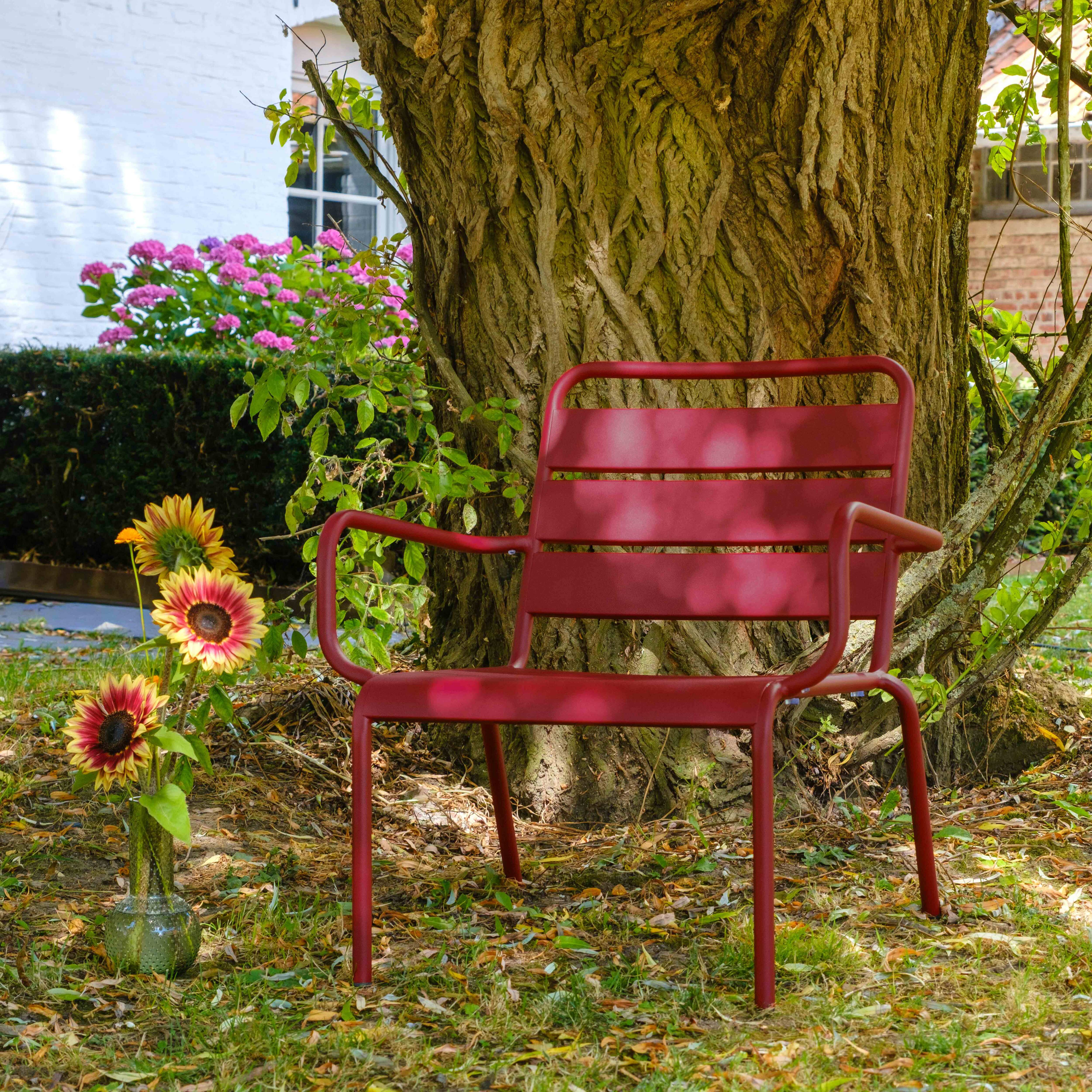 Ein roter Stuhl steht in der Nähe eines großen Baumes. Sonnenblumen stehen in einer Vase. Rosa Blumen und ein weißes Gebäude sind im Hintergrund.