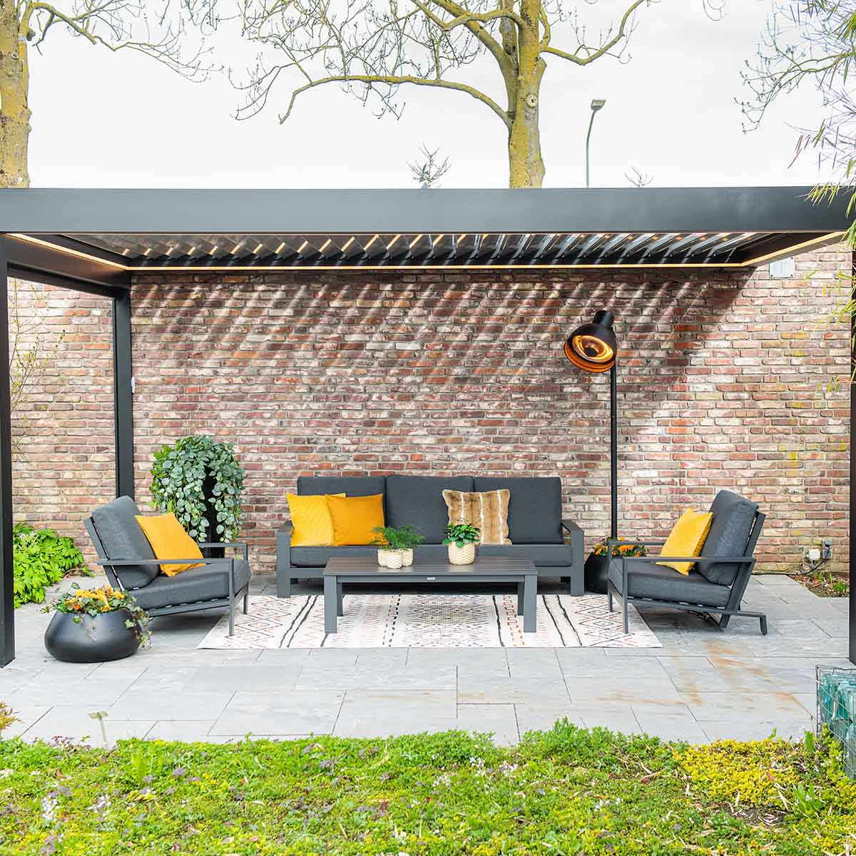 Outdoor patio with gray furniture, yellow pillows, rug, brick wall, and heater. Green grass and plants are in the foreground.