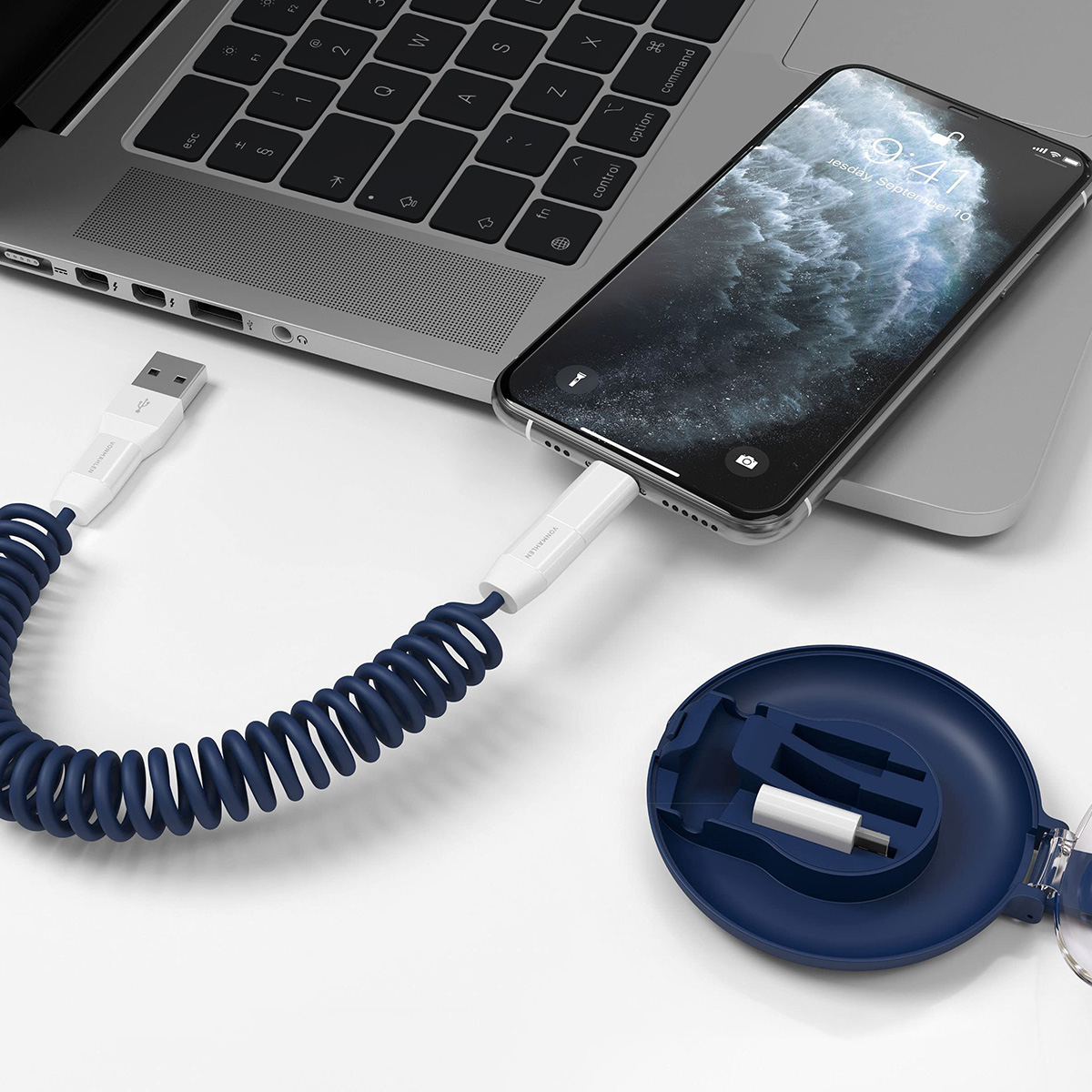 Un telefono è collegato a un laptop tramite un cavo di ricarica blu a spirale. A destra c'è una custodia blu scuro. Il telefono mostra l'ora.