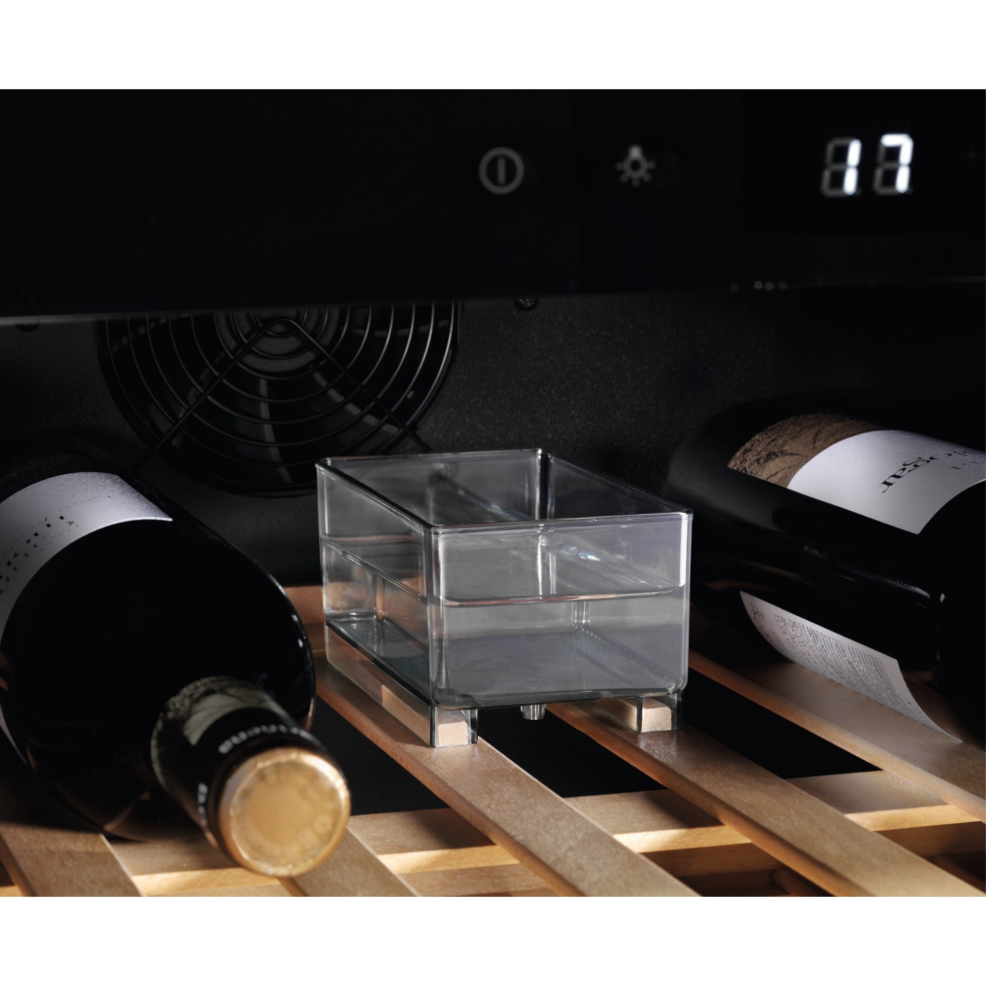 Dentro de una nevera para vinos, se ven dos botellas de vino, junto a un recipiente transparente. El fondo es oscuro.
